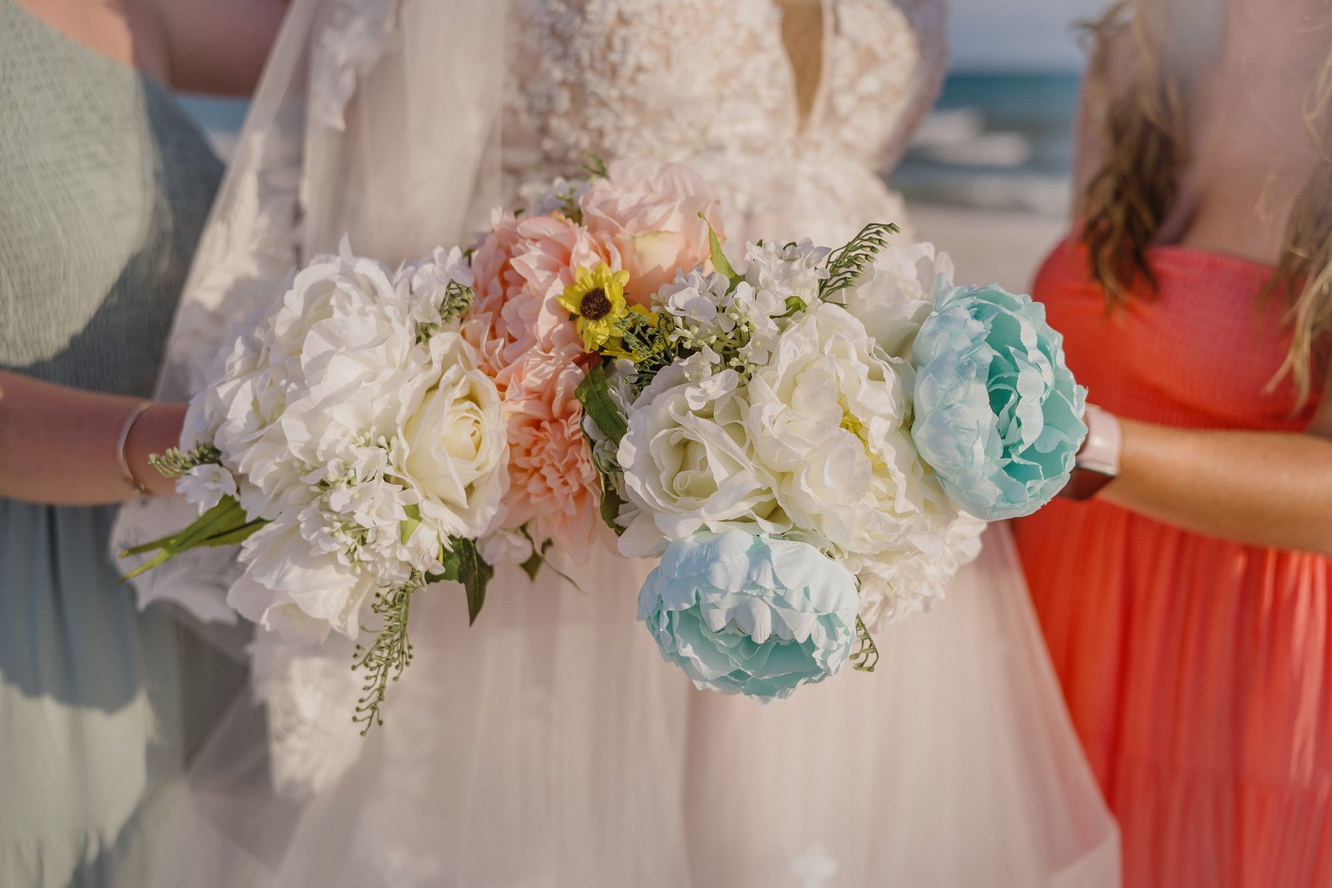 Bridal bouquet of white, blush, and blue flowers held against a wedding dress on a beach