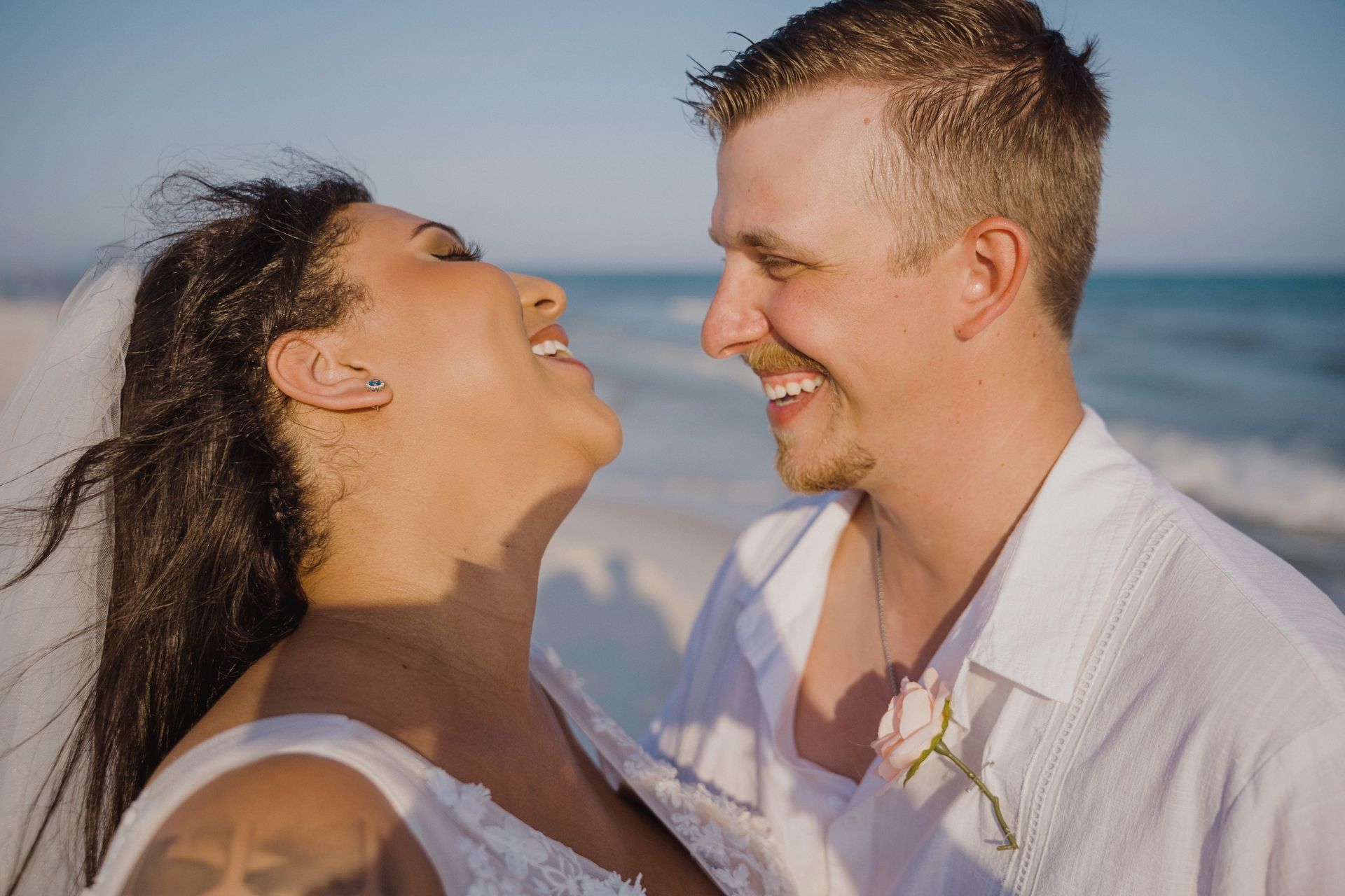 Smiling couple laughing together on a sunny beach, with the ocean blurred in the background