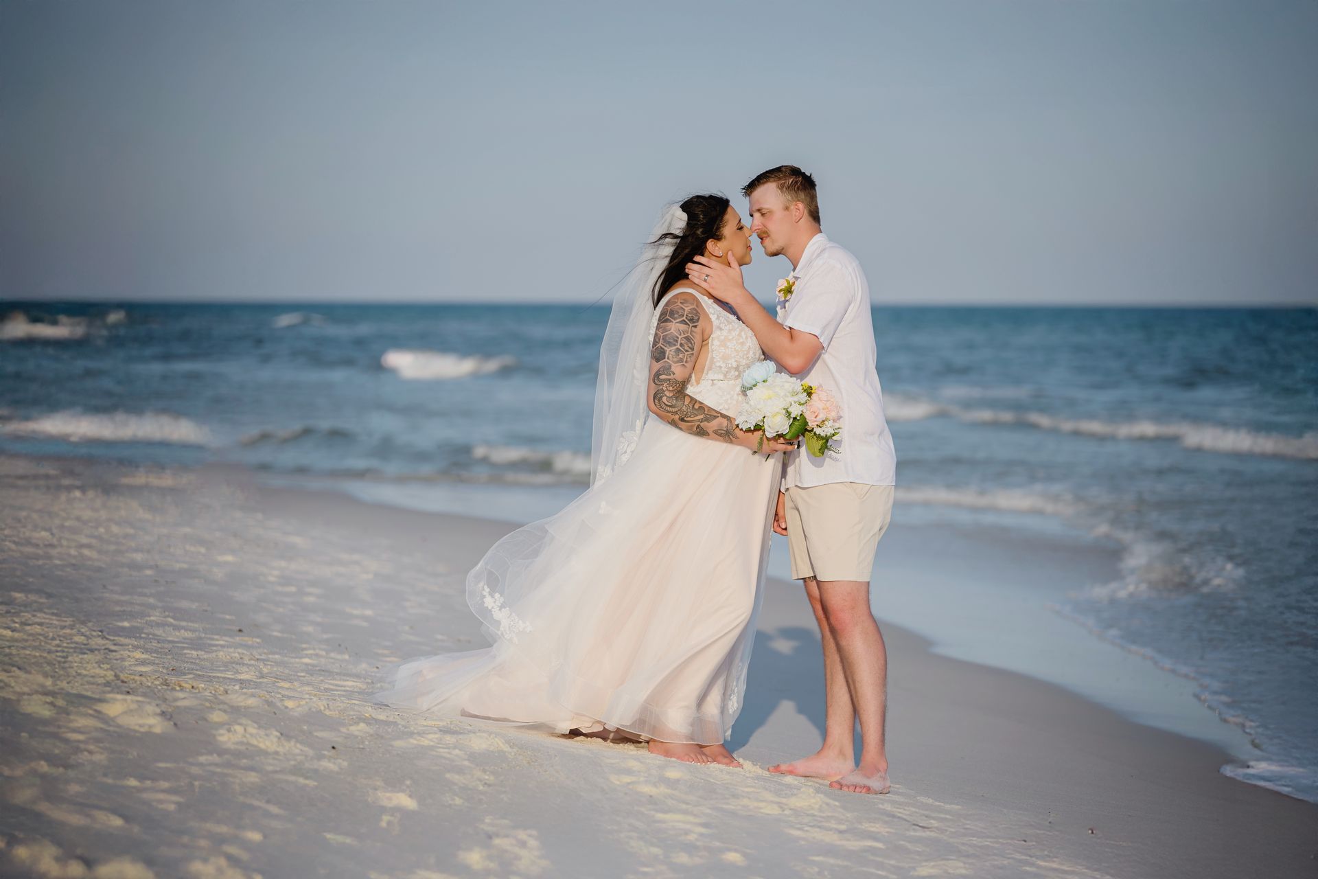 Bride and groom embrace on a beach at sunset, waves in the background.