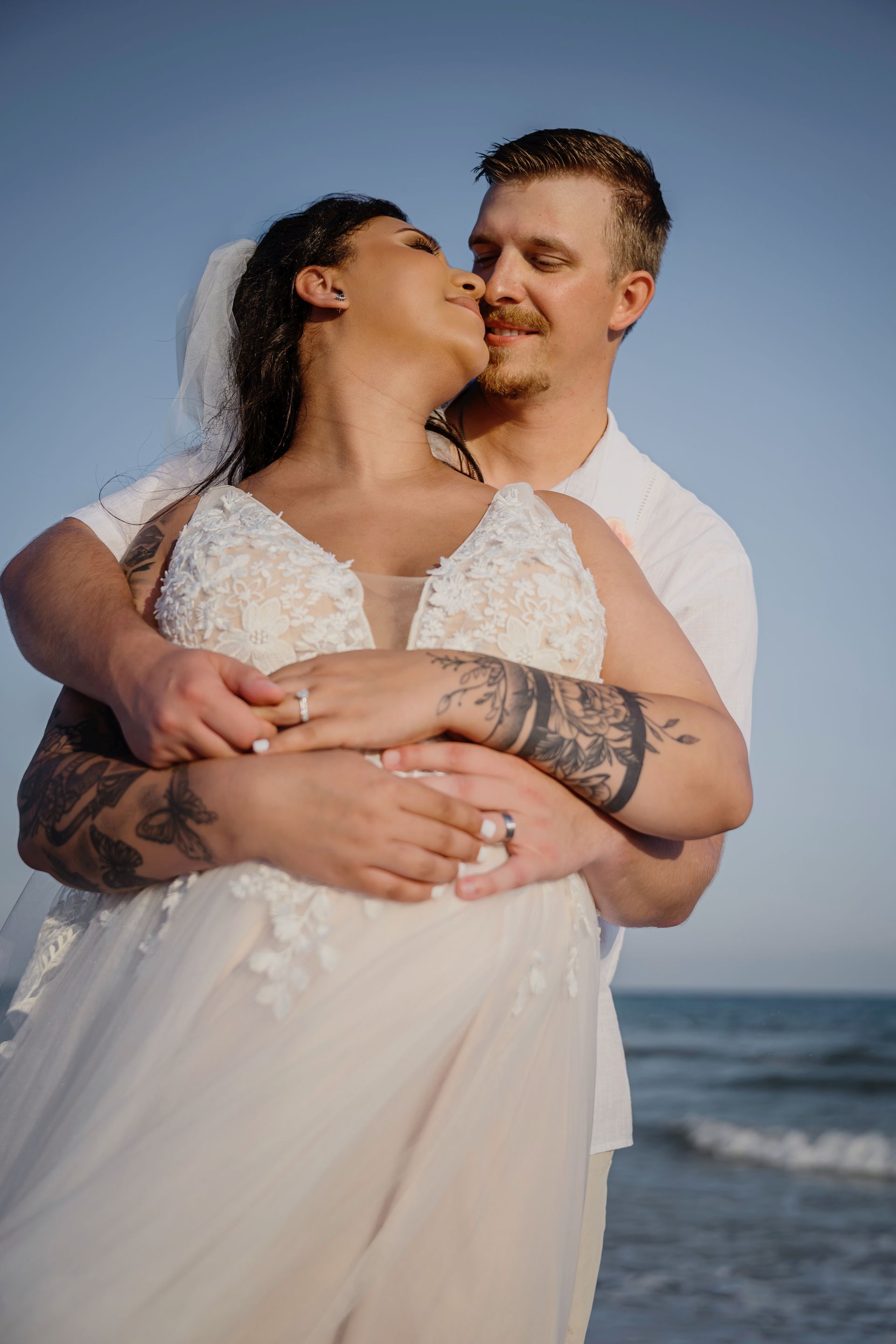 Couple embracing and kissing on a beach at sunset, with ocean waves behind them.
