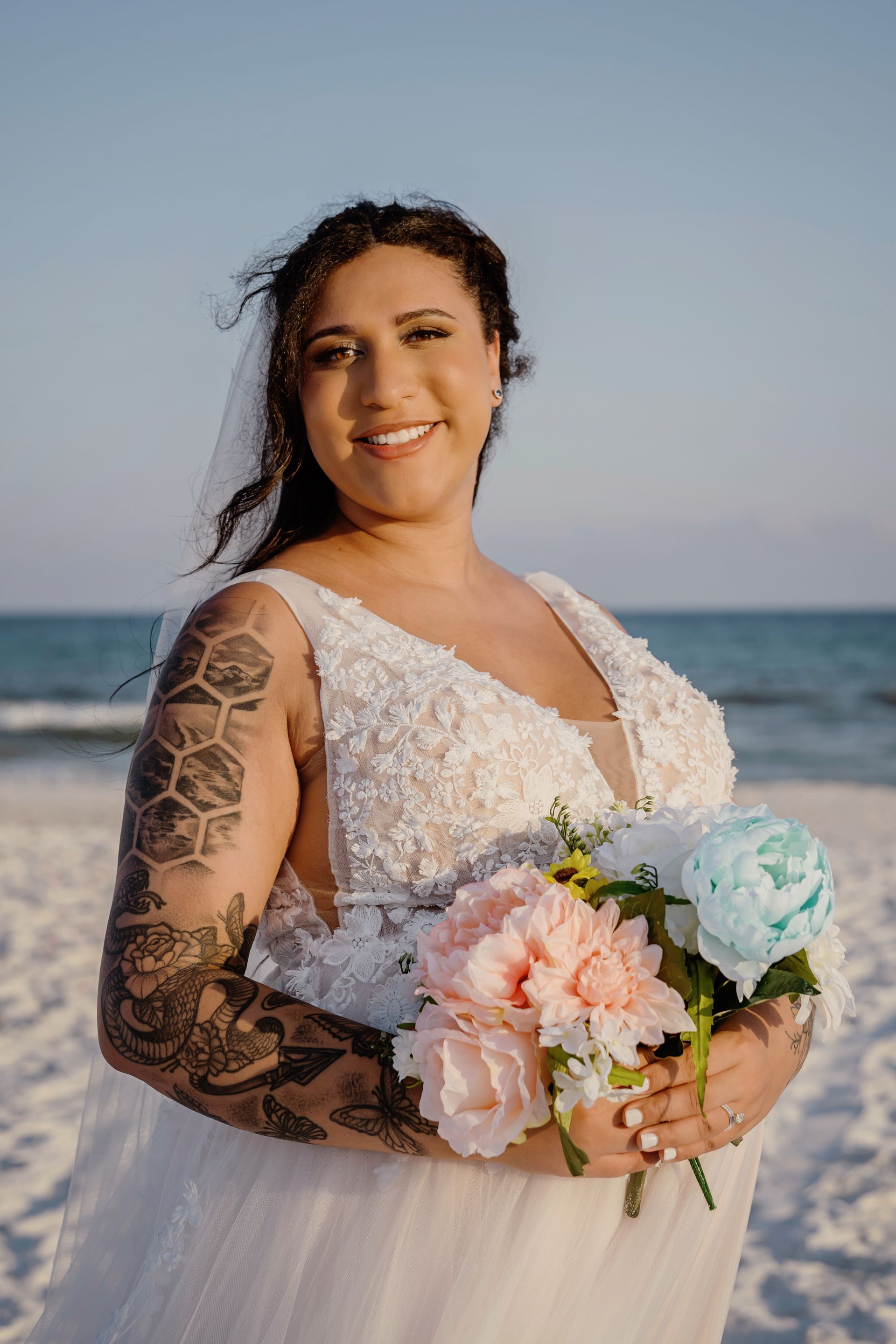 Woman in a white lace dress holding a pastel bouquet on a beach, smiling at sunset.