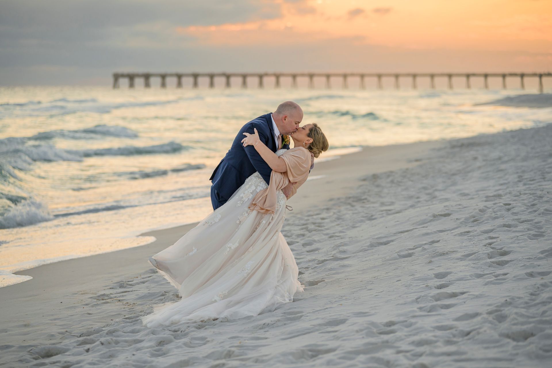 Couple embracing and dancing on a beach at sunset with waves and a pier in the background
