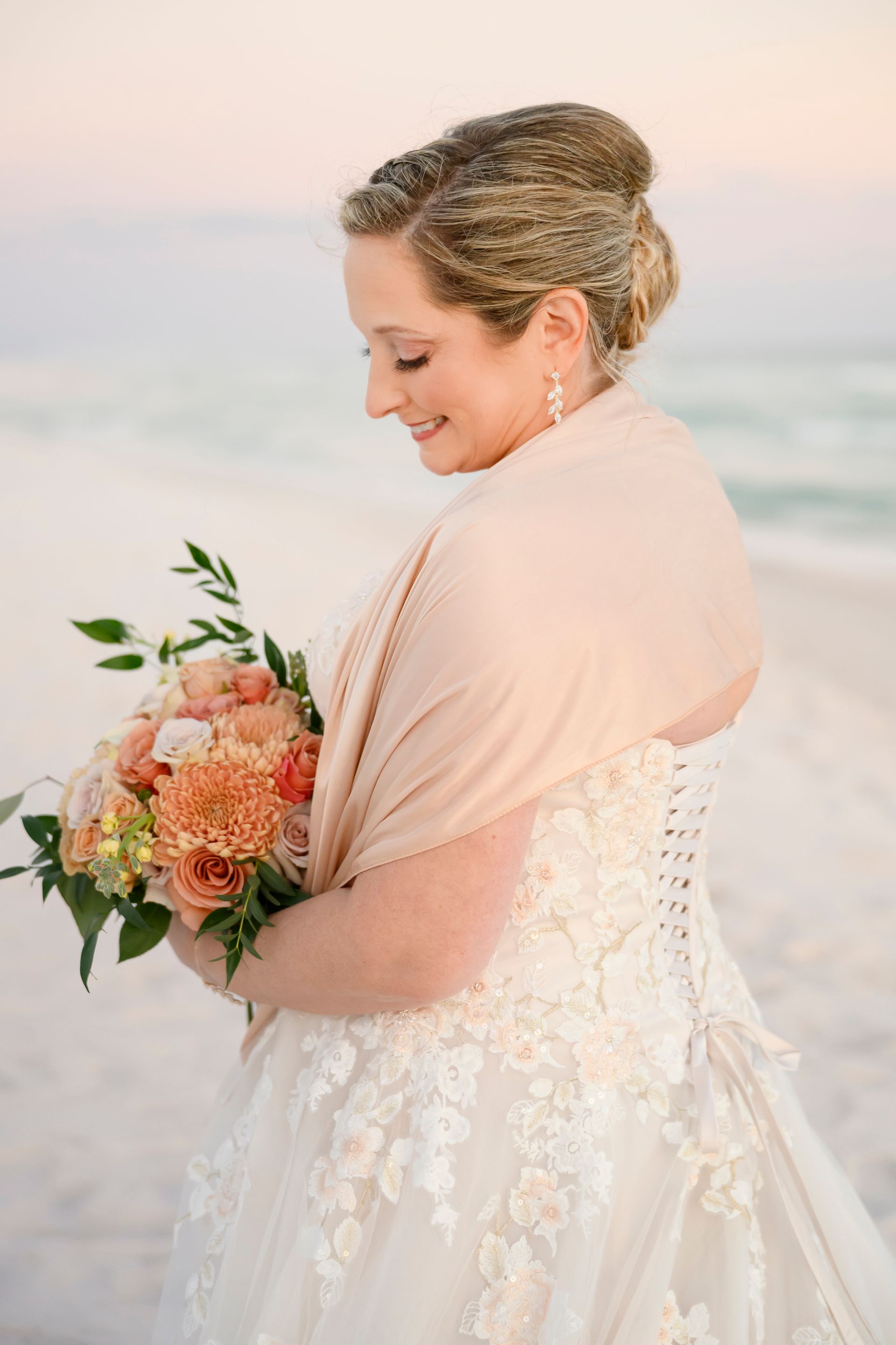Bride in a lace gown holding a peach and coral bouquet on a beach at sunset