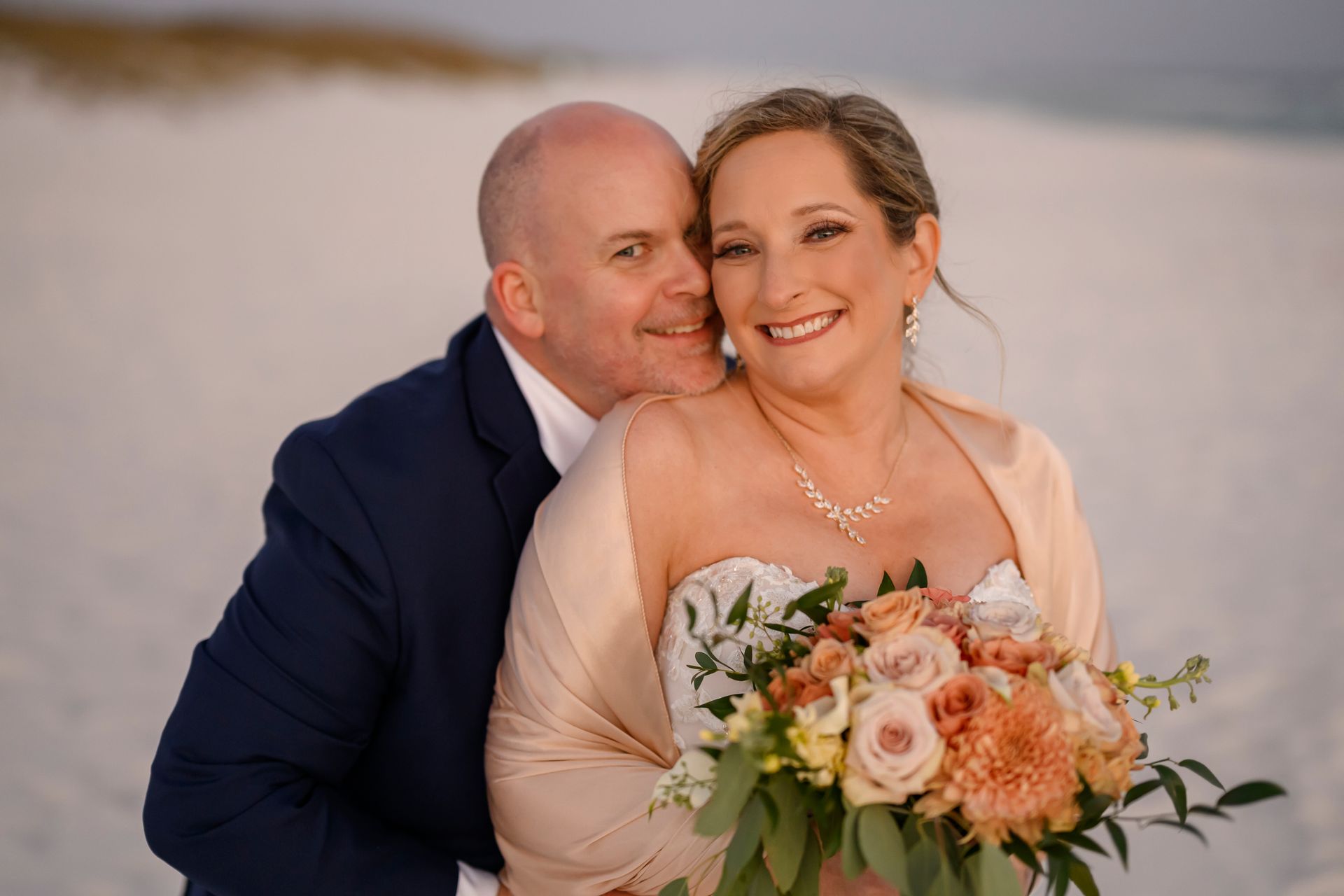 Couple embracing on a beach at sunset, smiling with a bouquet of peach and white flowers