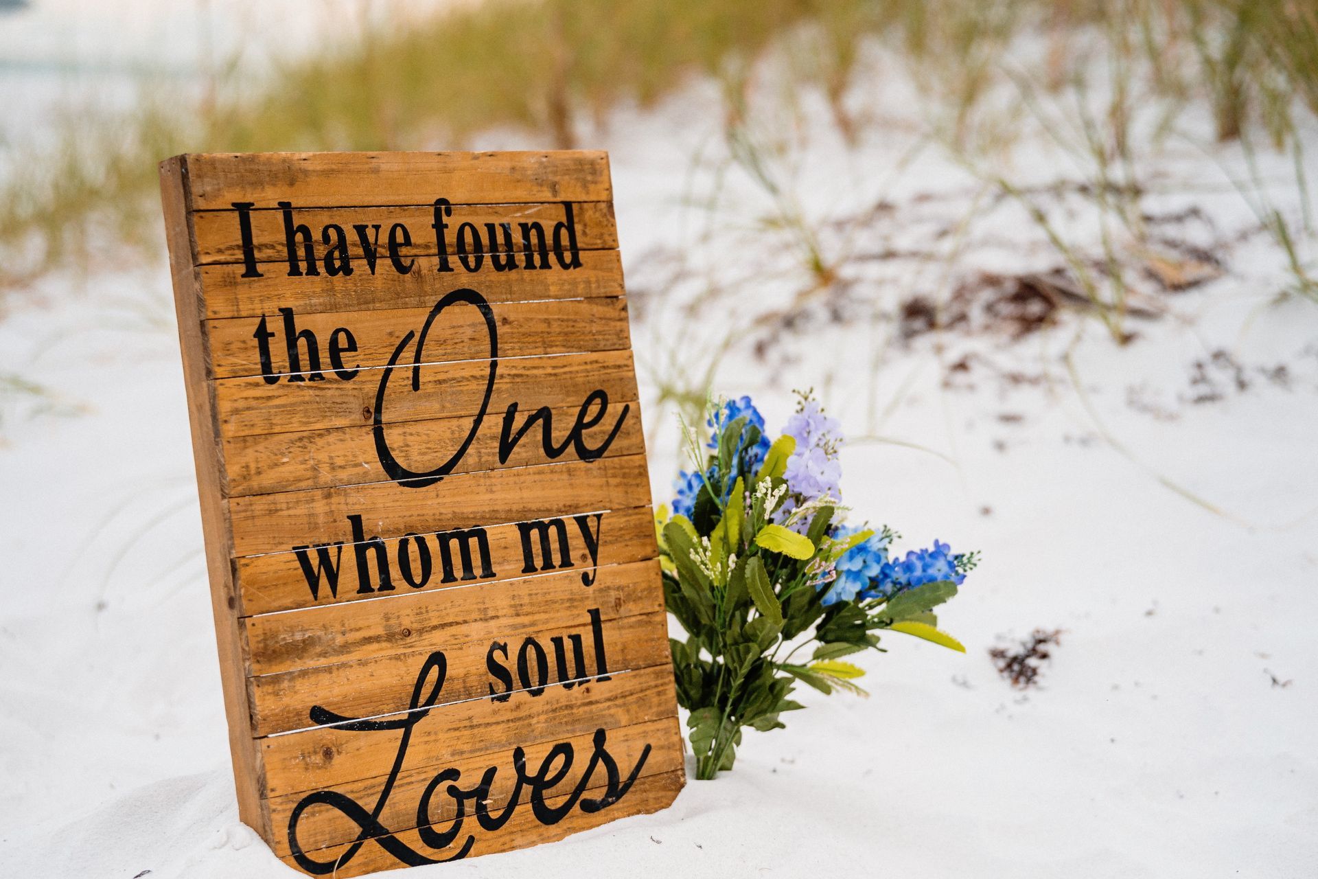 Wooden sign in snowy field reading “I have found the One whom my soul loves,” with blue-yellow flowers attached