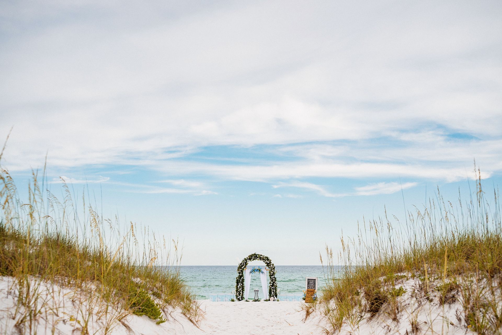 White sandy beach with tall grass framing a blue ocean and a flowered arch under a bright sky