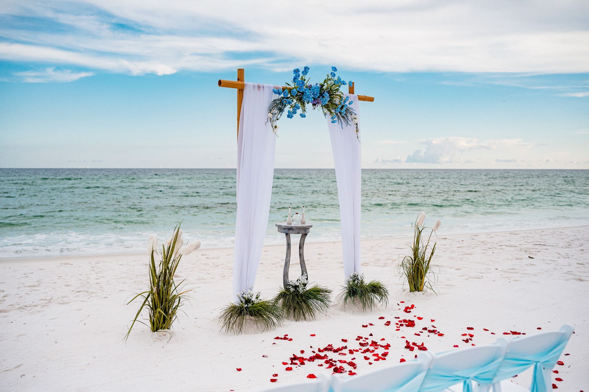 Beach wedding arch with white drapes, blue flowers, and red petals by the sea