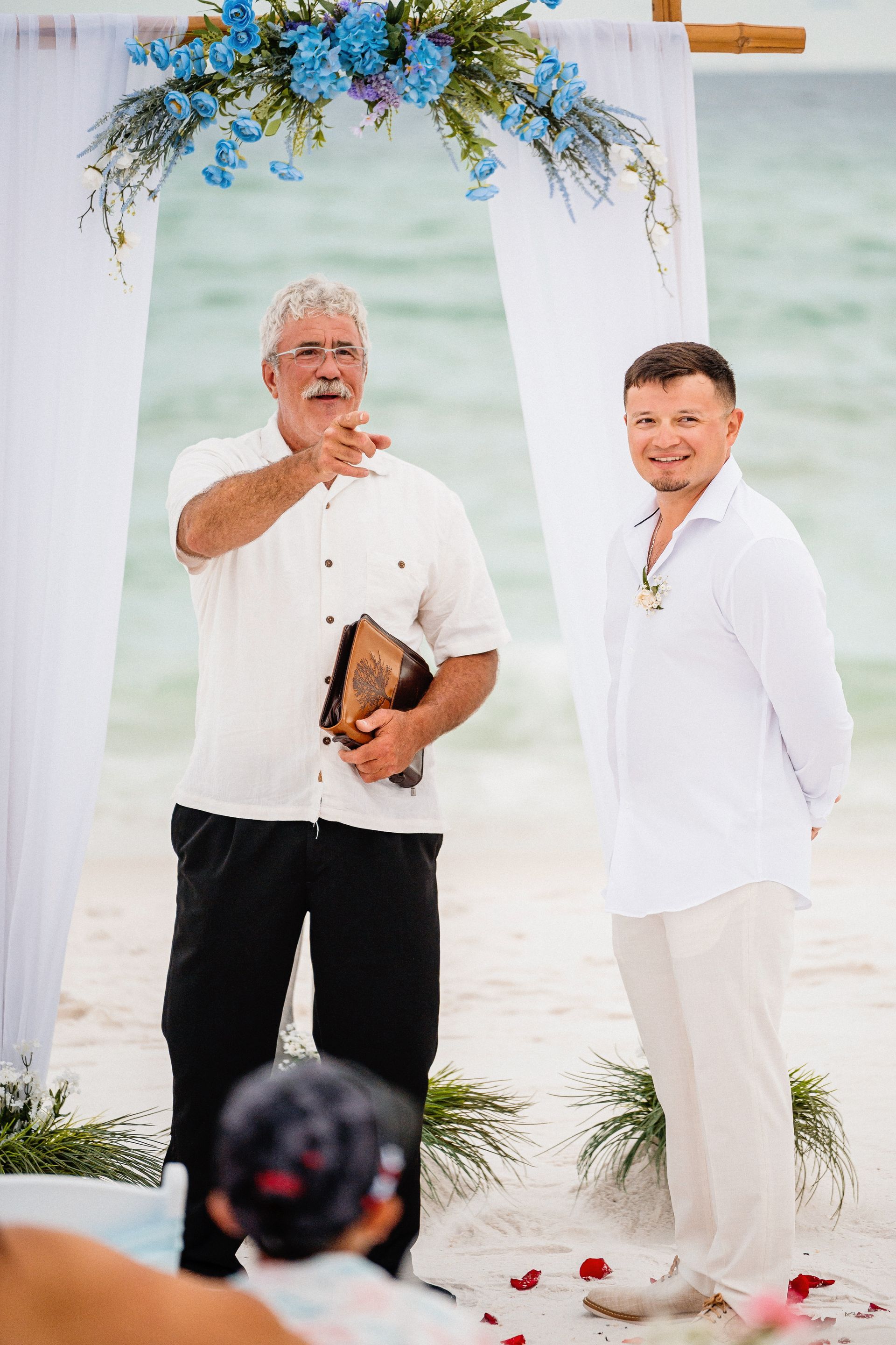 Two men standing under a floral arch on a beach, one holding a book and speaking into a microphone.