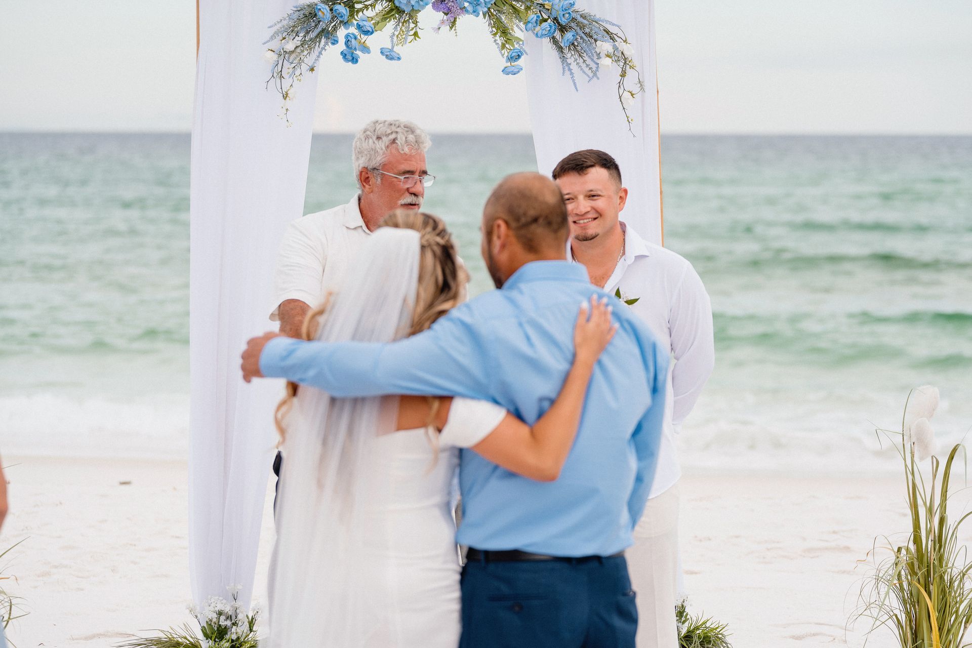 Four people embracing under a white arch on a beach with ocean waves behind them