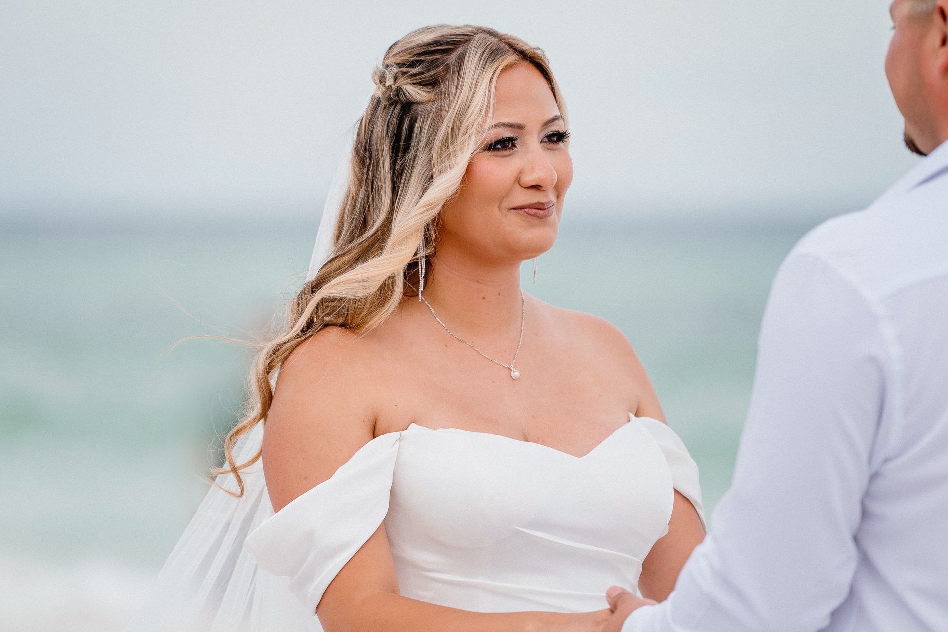 Bride in white dress smiling at a man on a beach with the ocean behind her