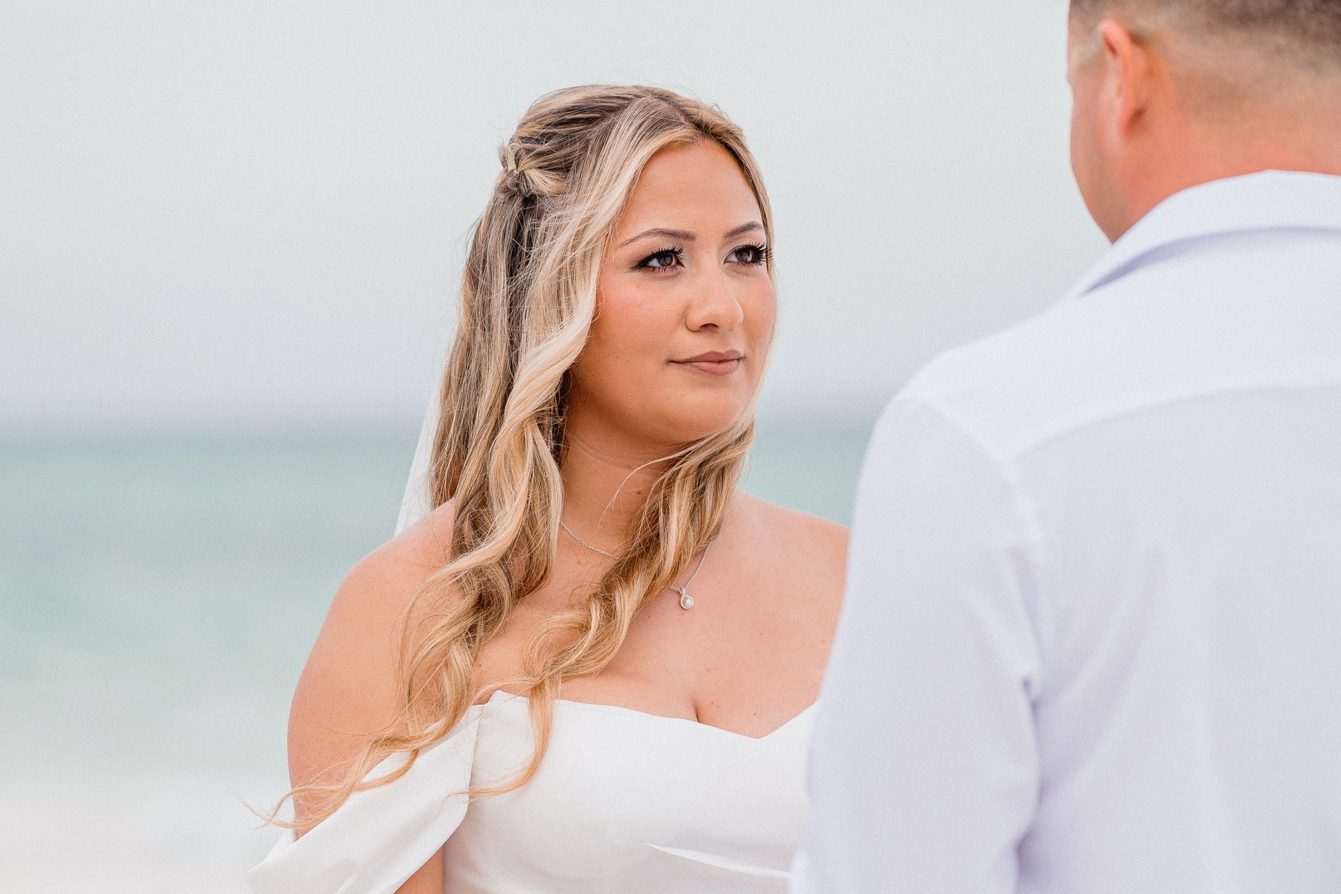 Bride in a white dress faces a man in a white shirt at a beach ceremony