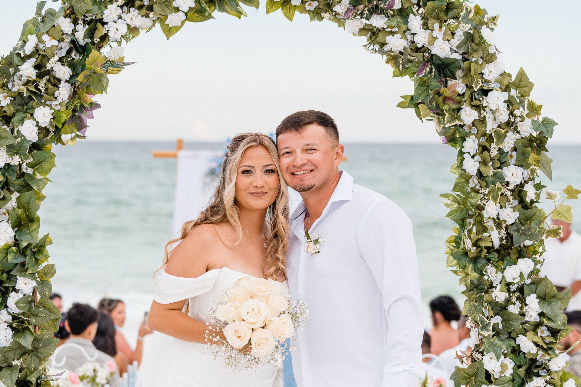 Couple in white wedding attire posing under a floral arch by the sea