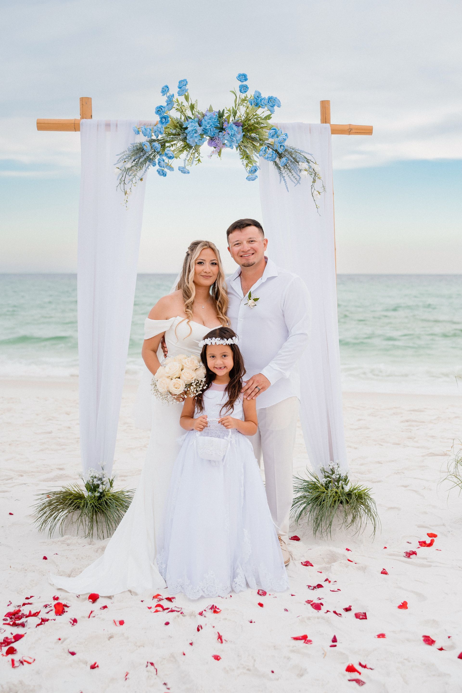 Family portrait under white floral arch on a beach, with ocean and rose petals in the sand.