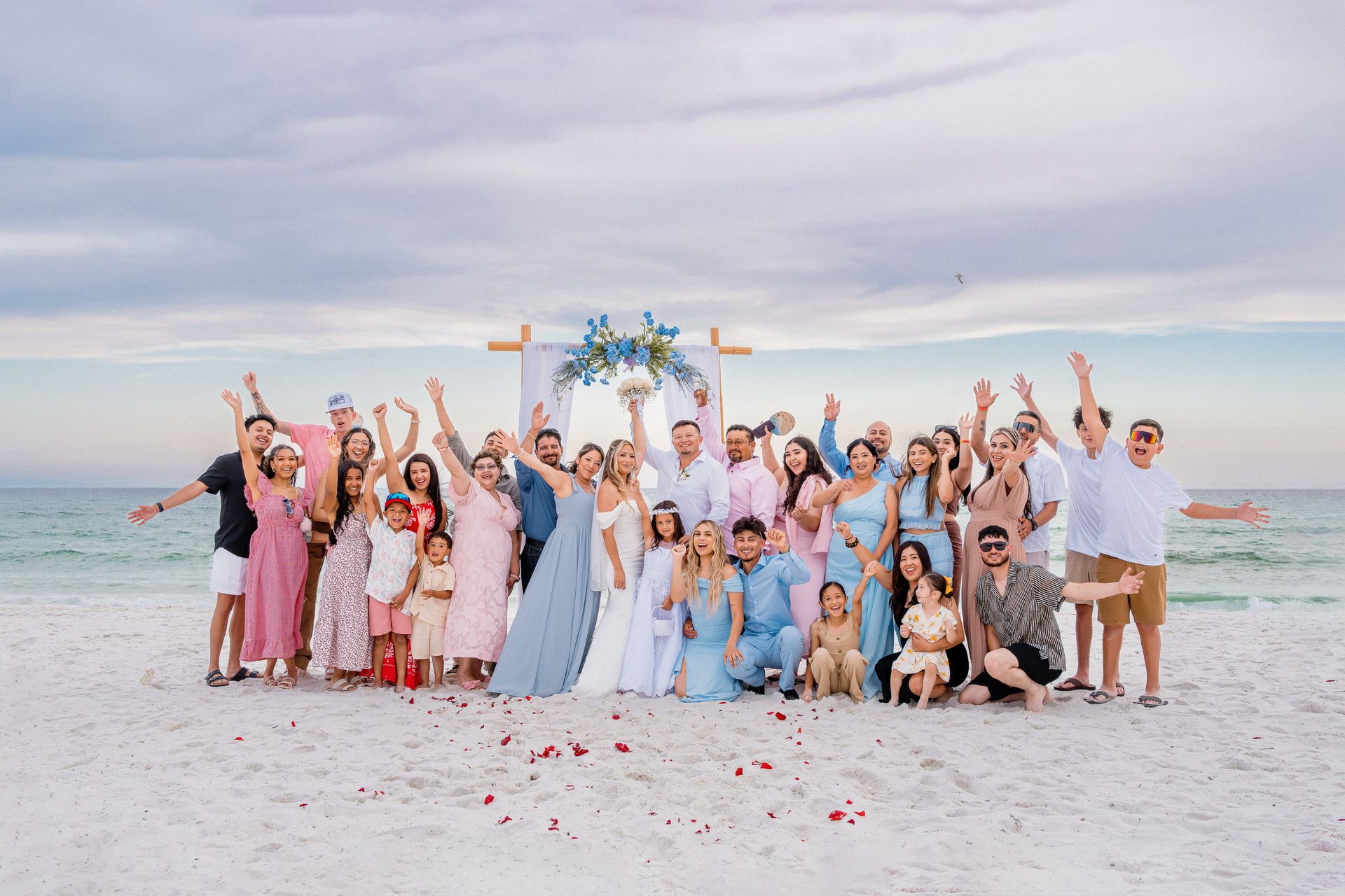 Group celebrating on a beach under a floral arch, with people raising their hands and confetti in the air.