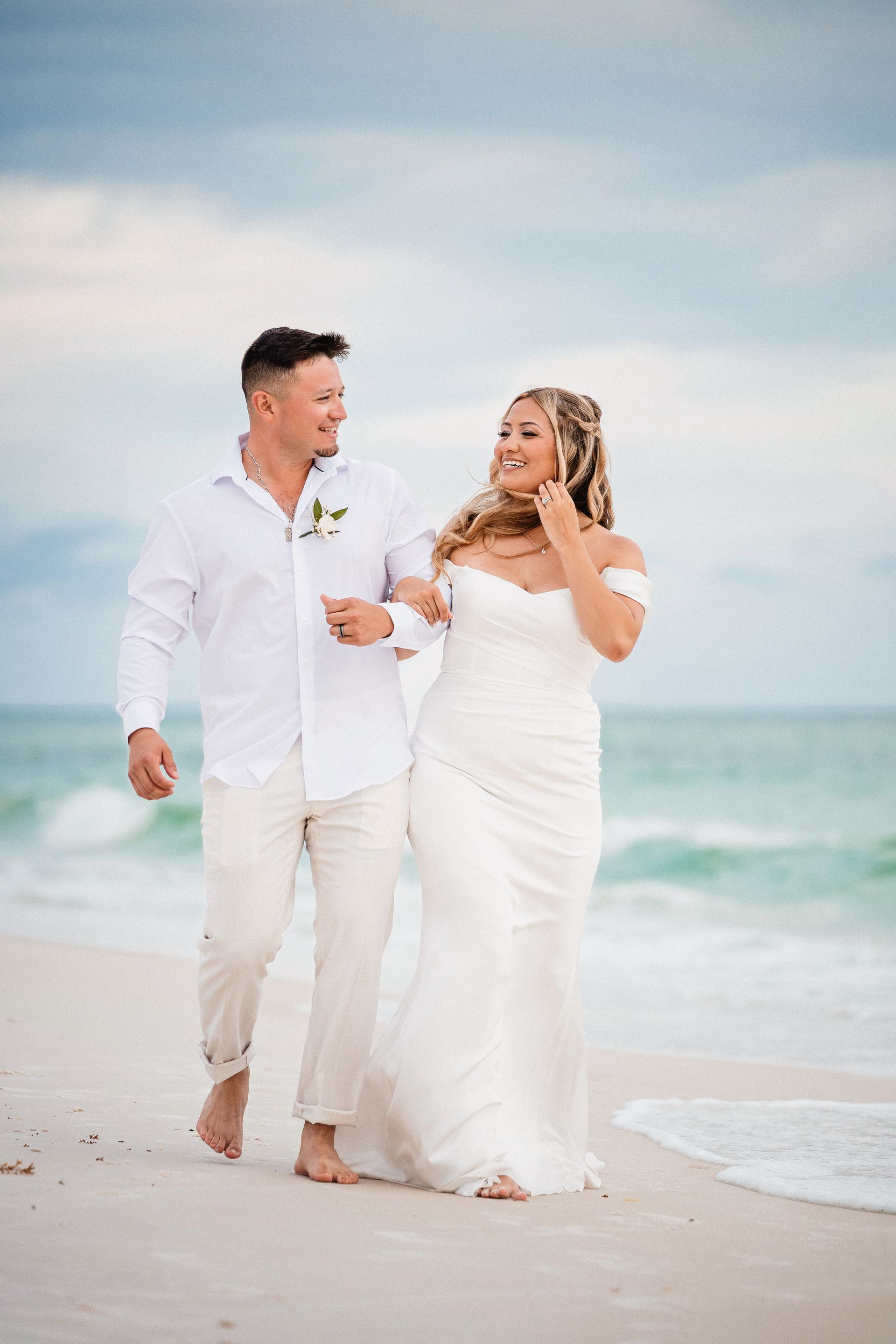 Couple in white walking barefoot on a beach, smiling and holding hands near the ocean