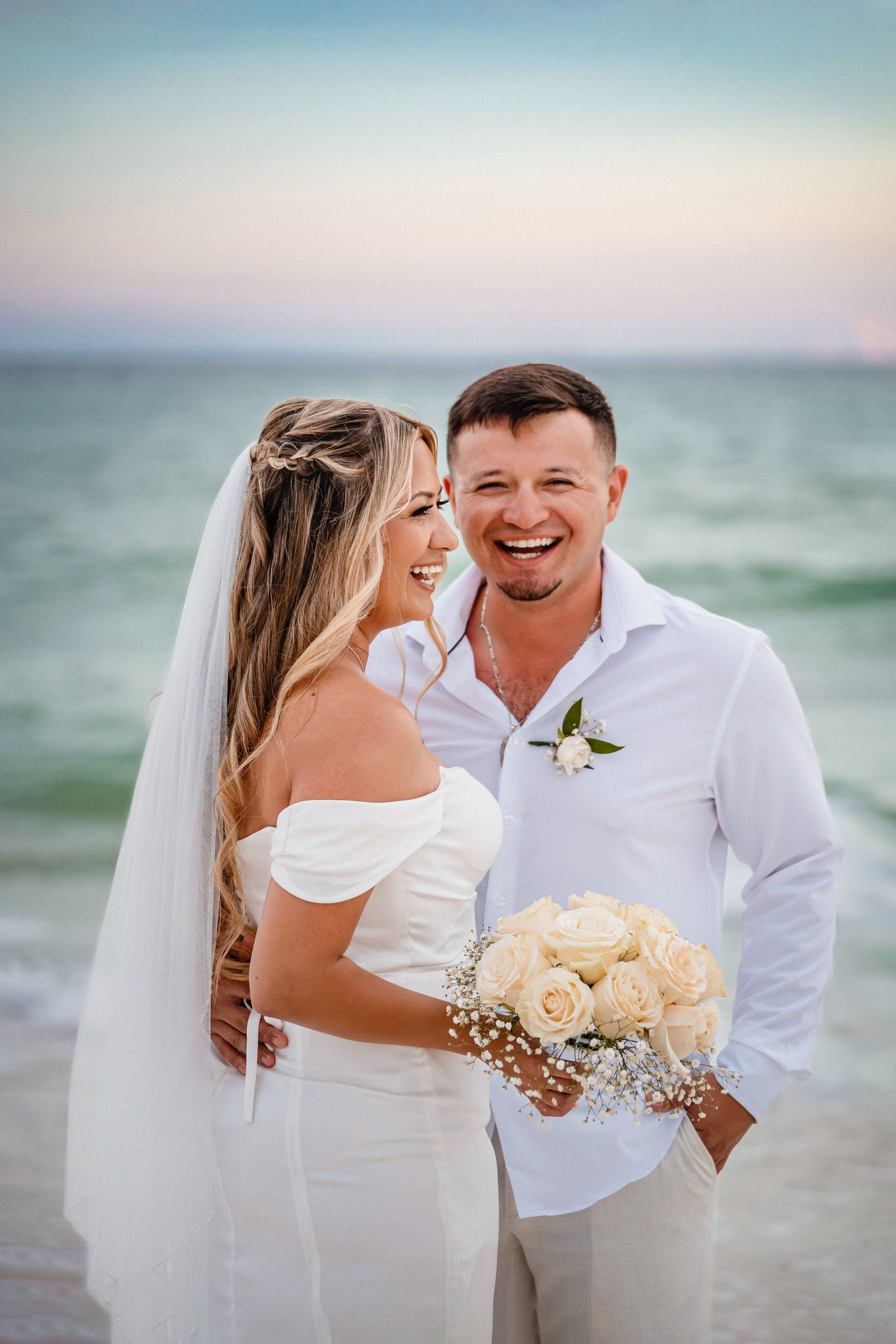 Couple in white wedding attire smiling on a beach, holding a bouquet by the ocean.