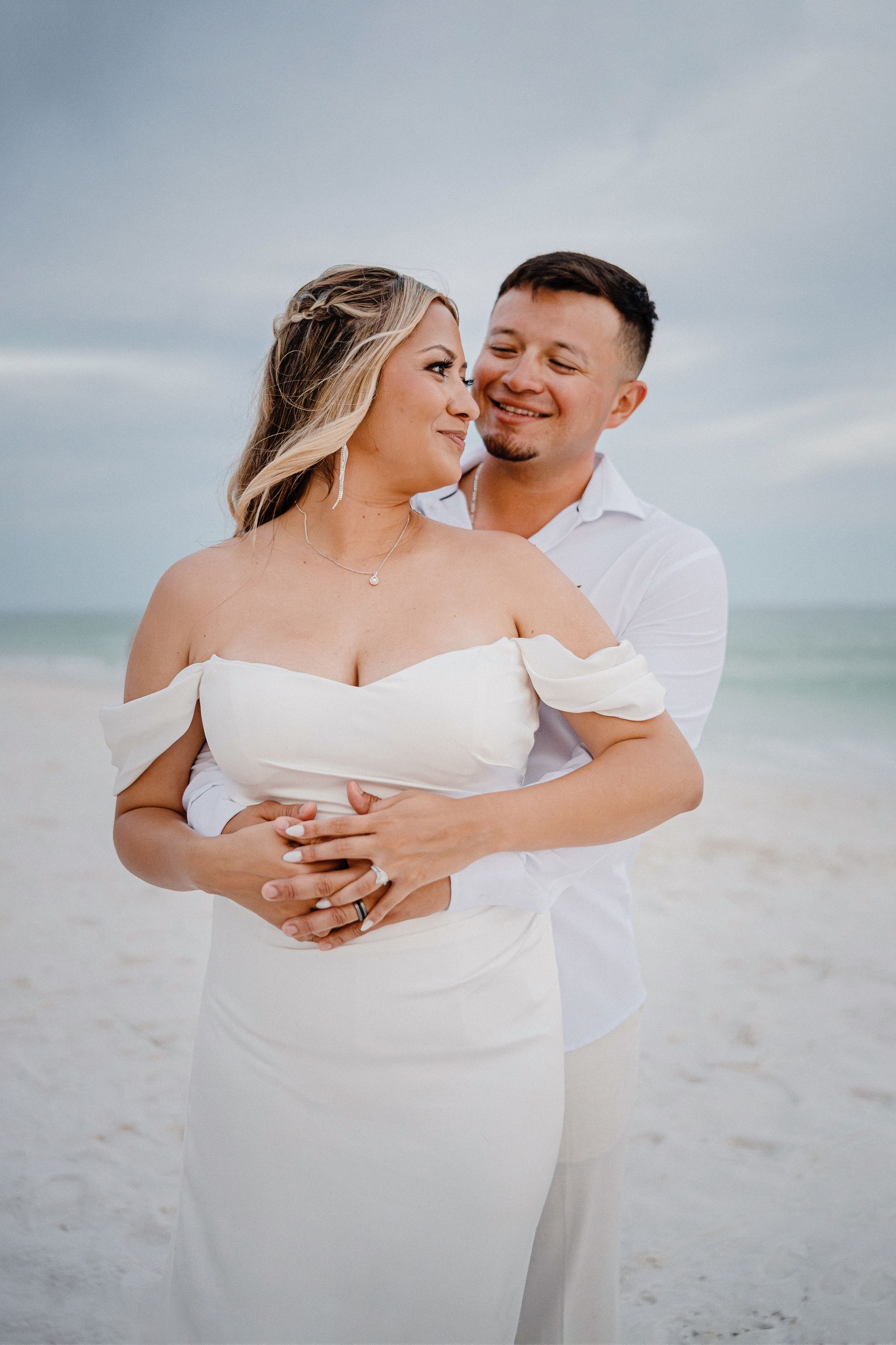 Couple embracing on a white sand beach at sunset, with the woman in a white dress and the man in a white shirt