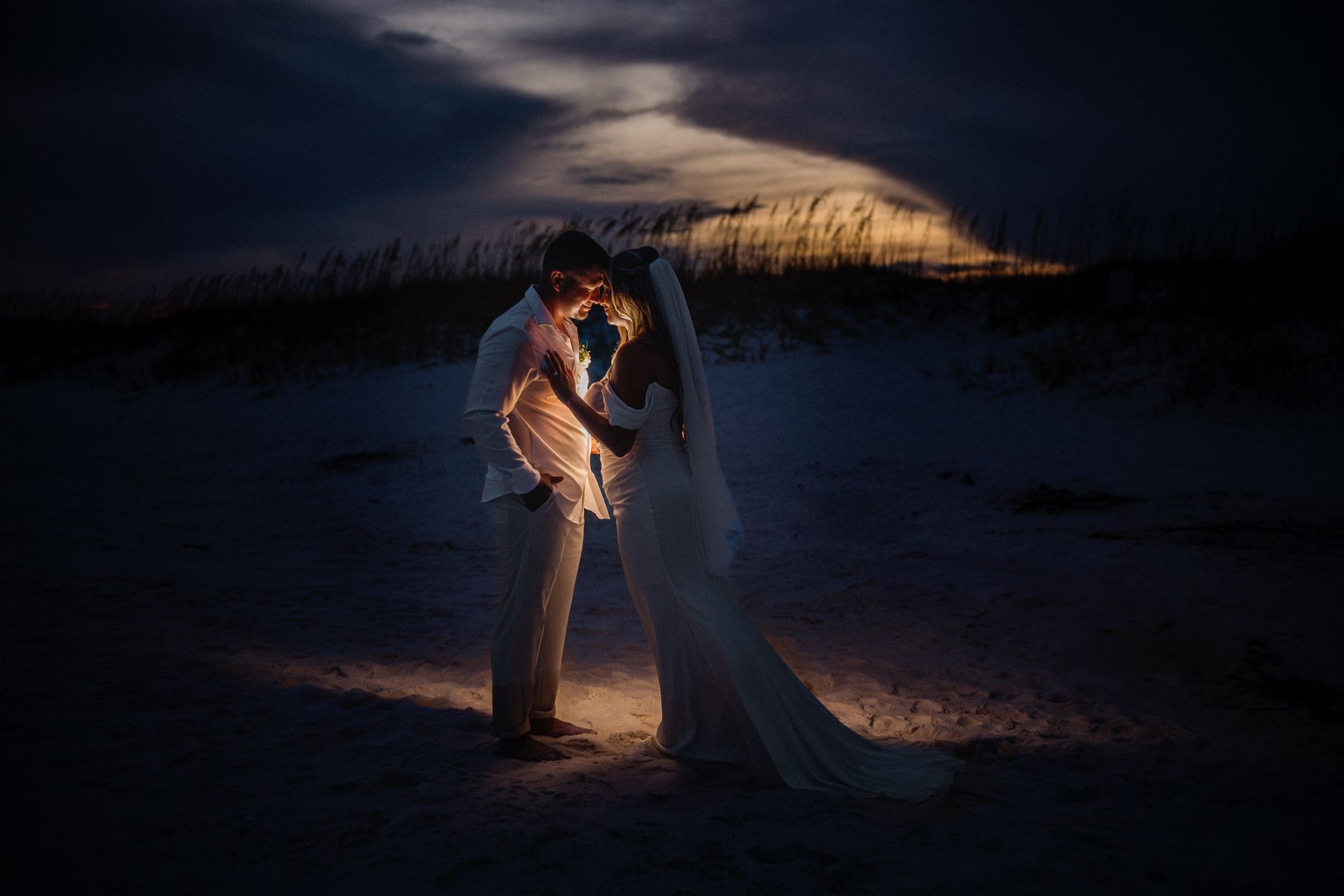 Couple embracing on a dark beach at dusk, lit by a warm glow against a cloudy sky