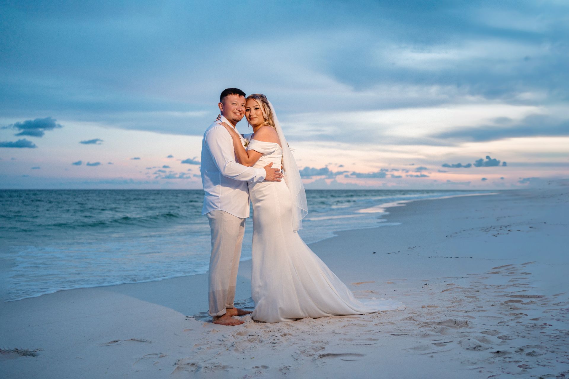 Couple in white wedding attire embracing on a beach at sunset with calm ocean waves.