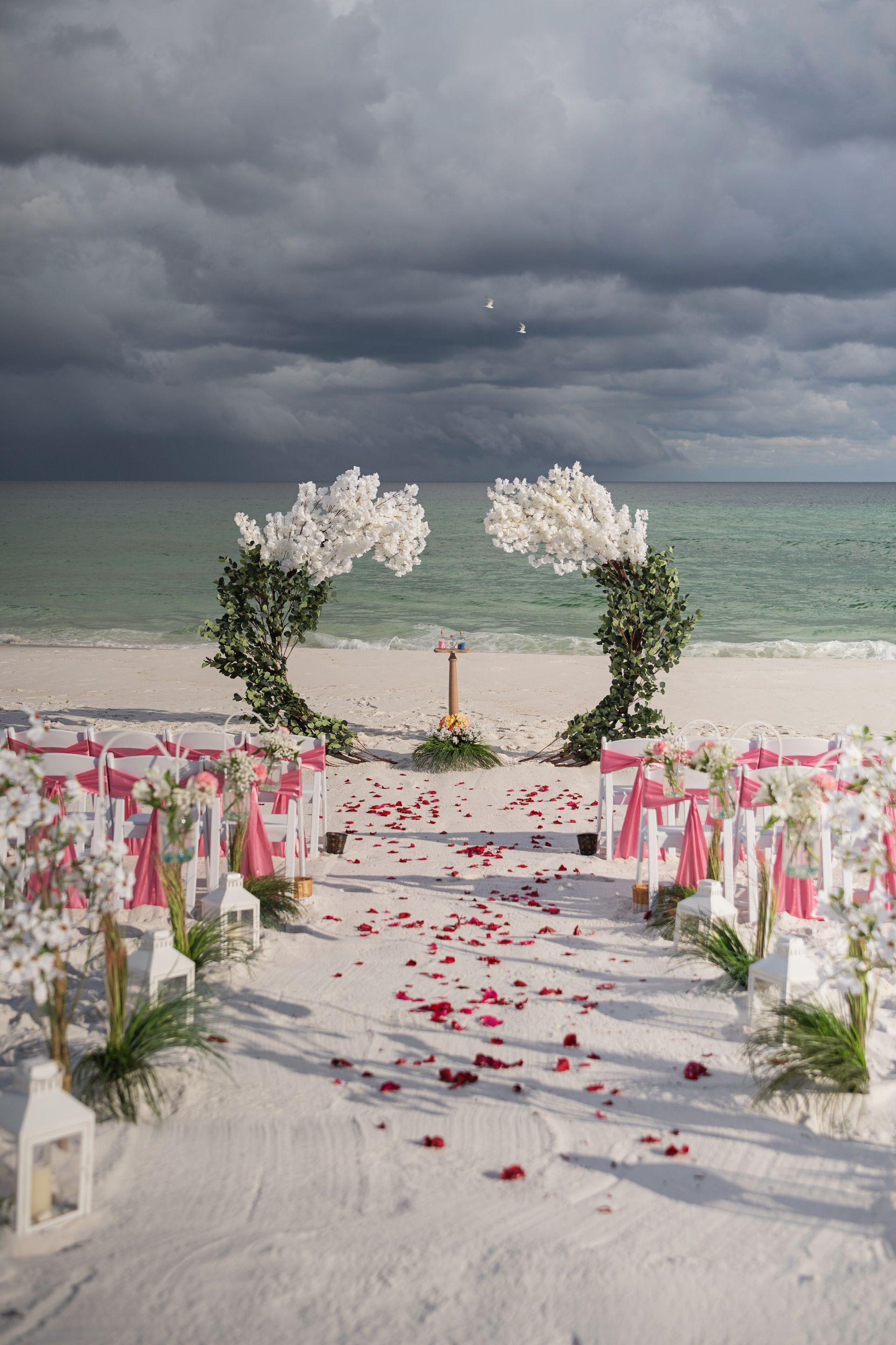 Beach wedding aisle with white flowers, pink petals, and ocean under dark clouds