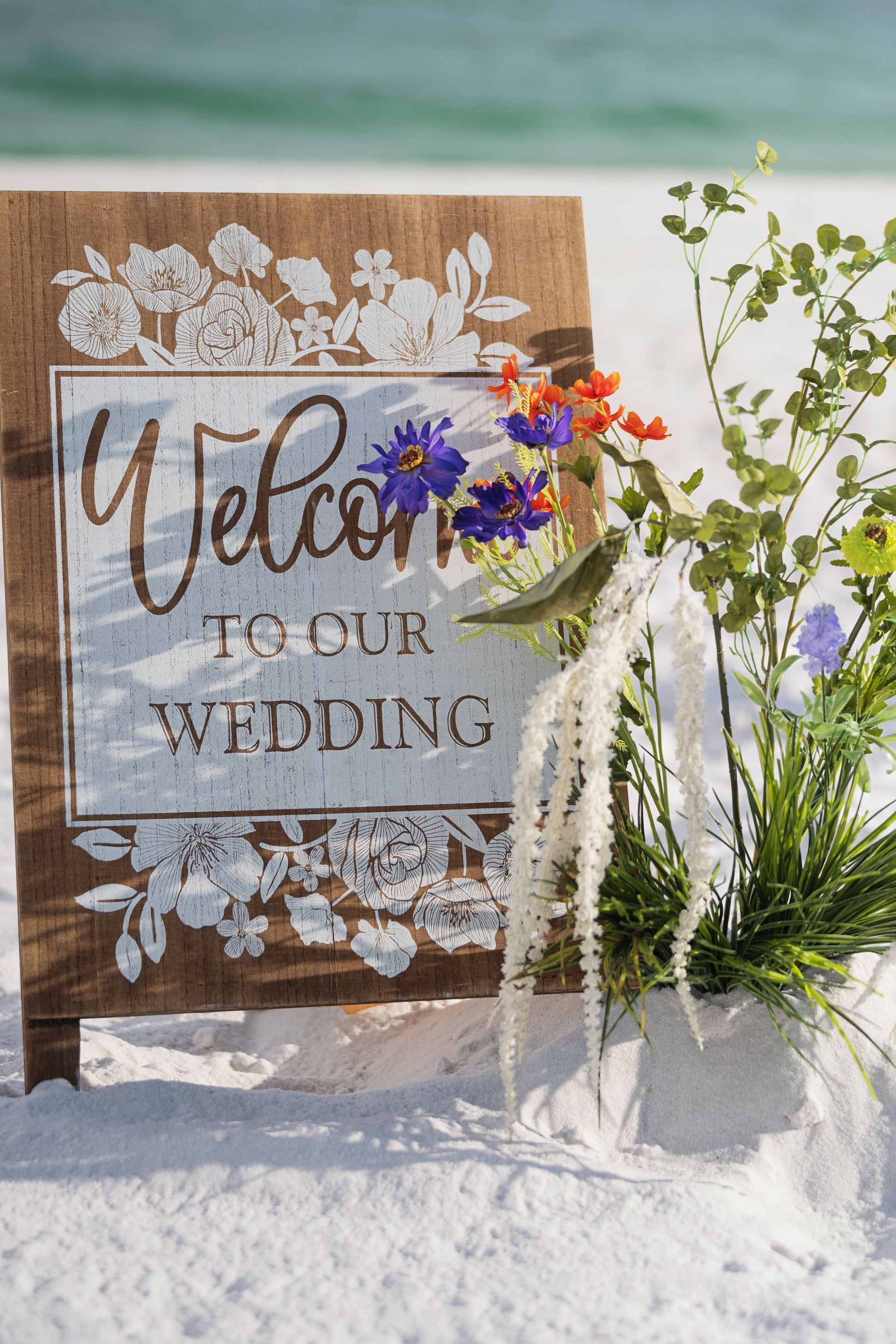 Beach wedding welcome sign with white floral decor and purple flowers in sand