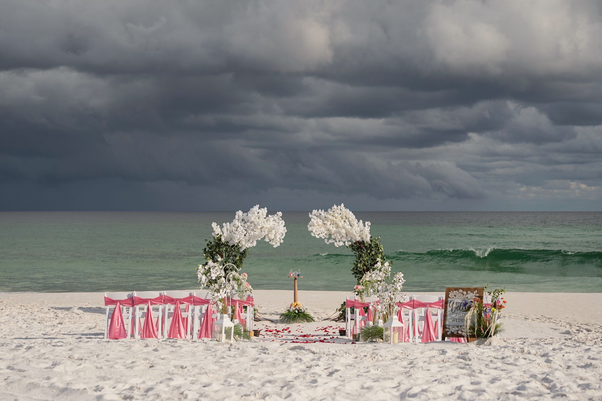 Beach wedding setup with pink chairs and white floral arch under stormy clouds