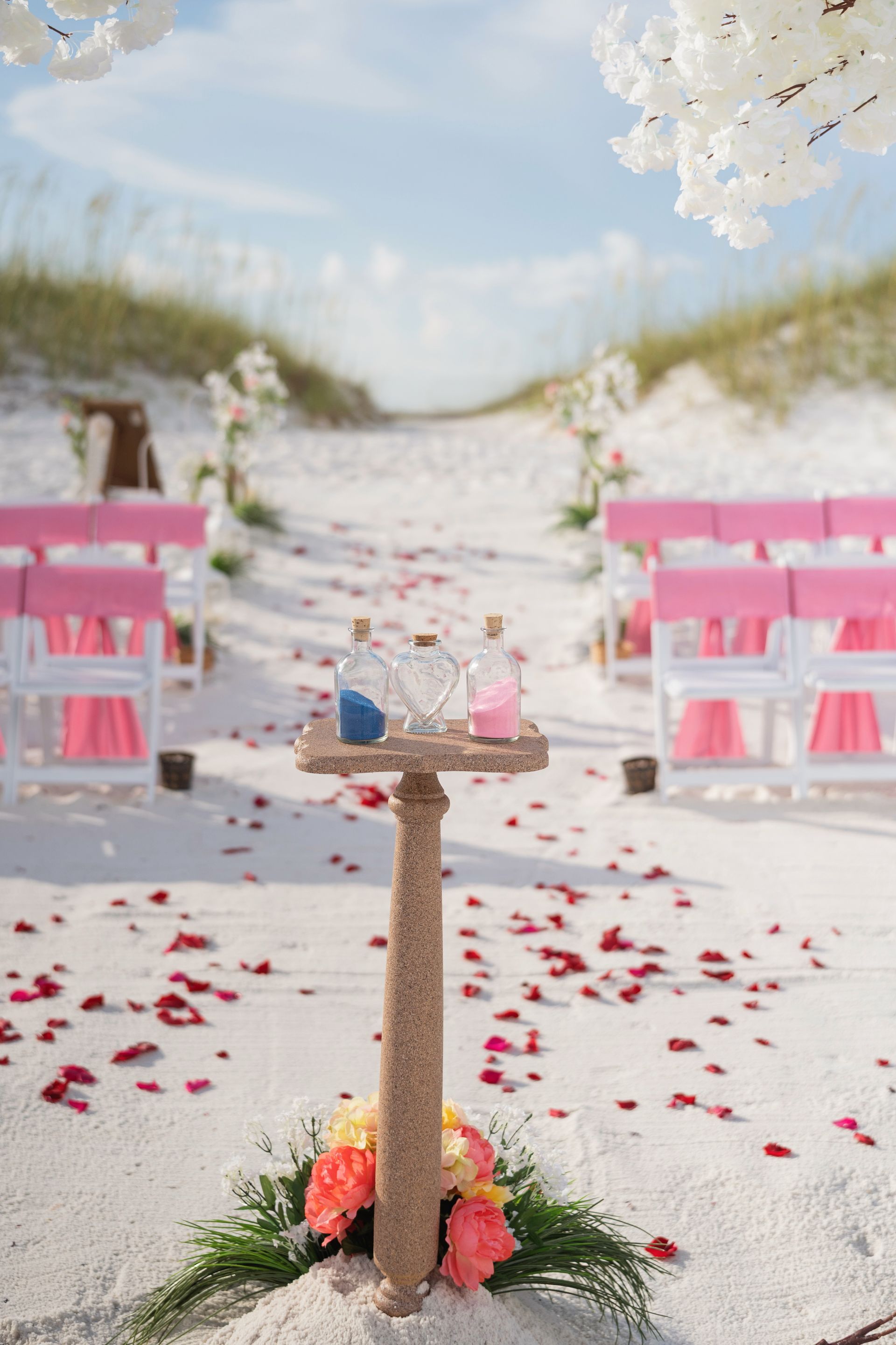 Beach wedding aisle with pink chairs, a small sand-colored pedestal, and scattered rose petals under white flowers