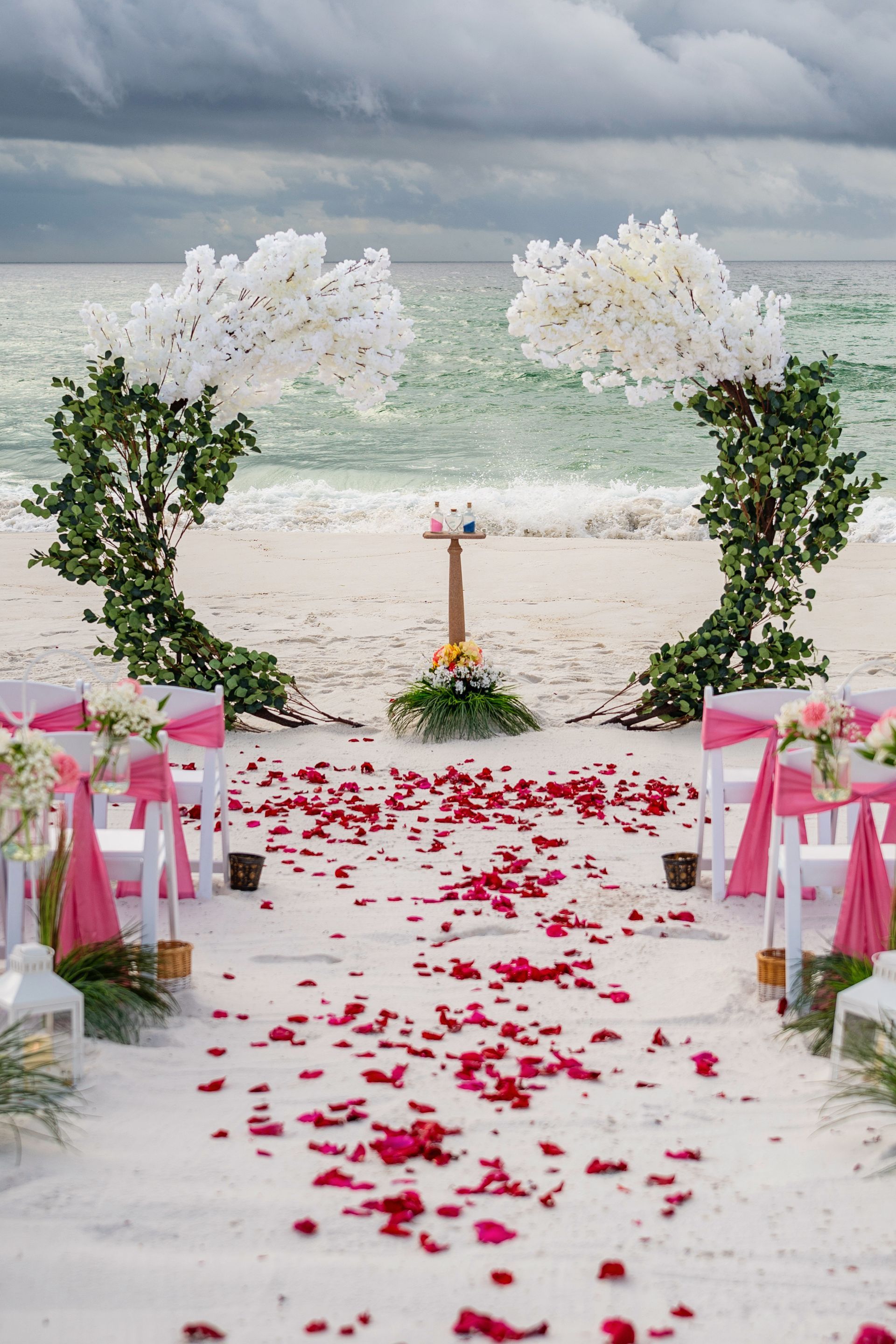 Beach wedding aisle with white floral arch, pink chairs, and red rose petals on sand under cloudy sky