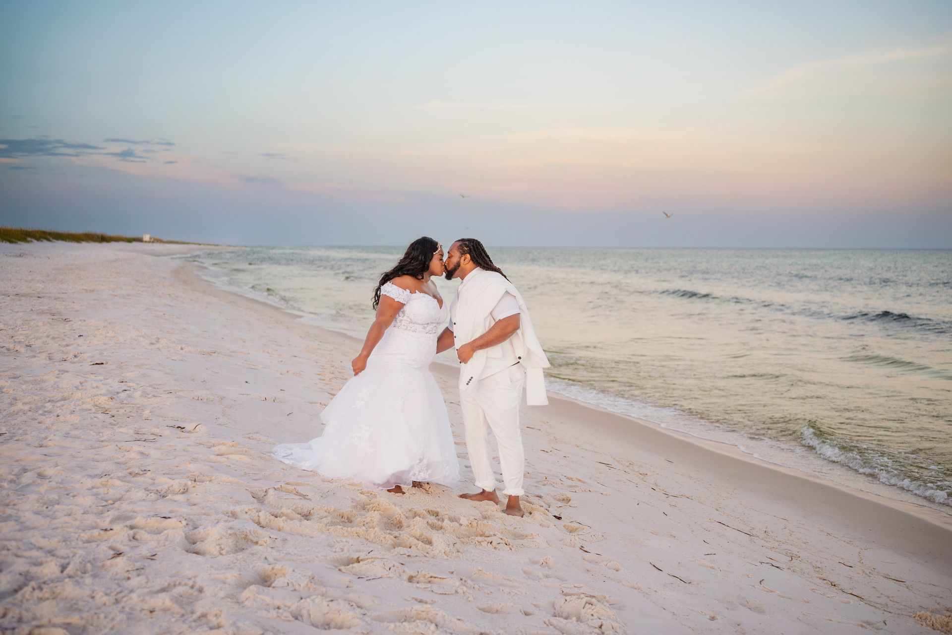 Three people in white outfits embracing on a sandy beach at sunset