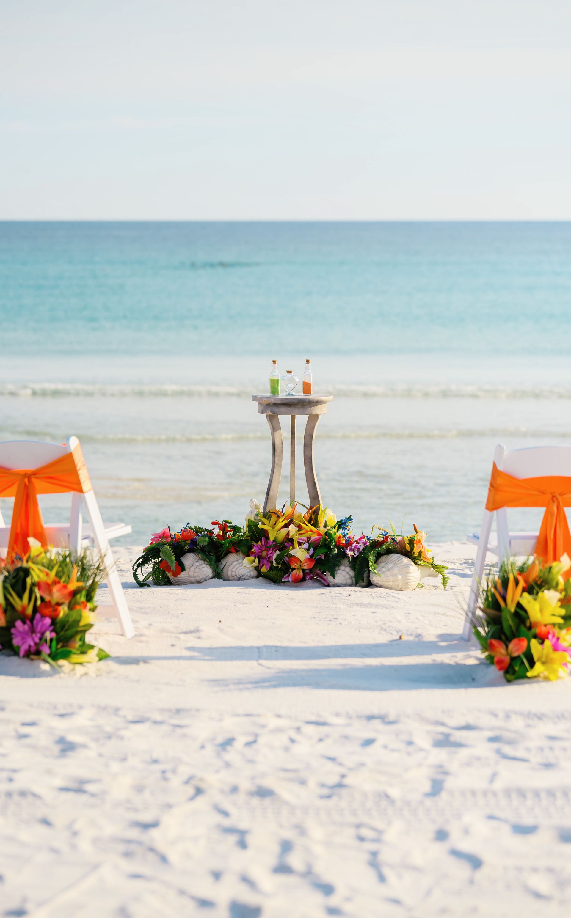 Beach wedding arch with colorful flowers, lanterns, and orange chairs facing the ocean