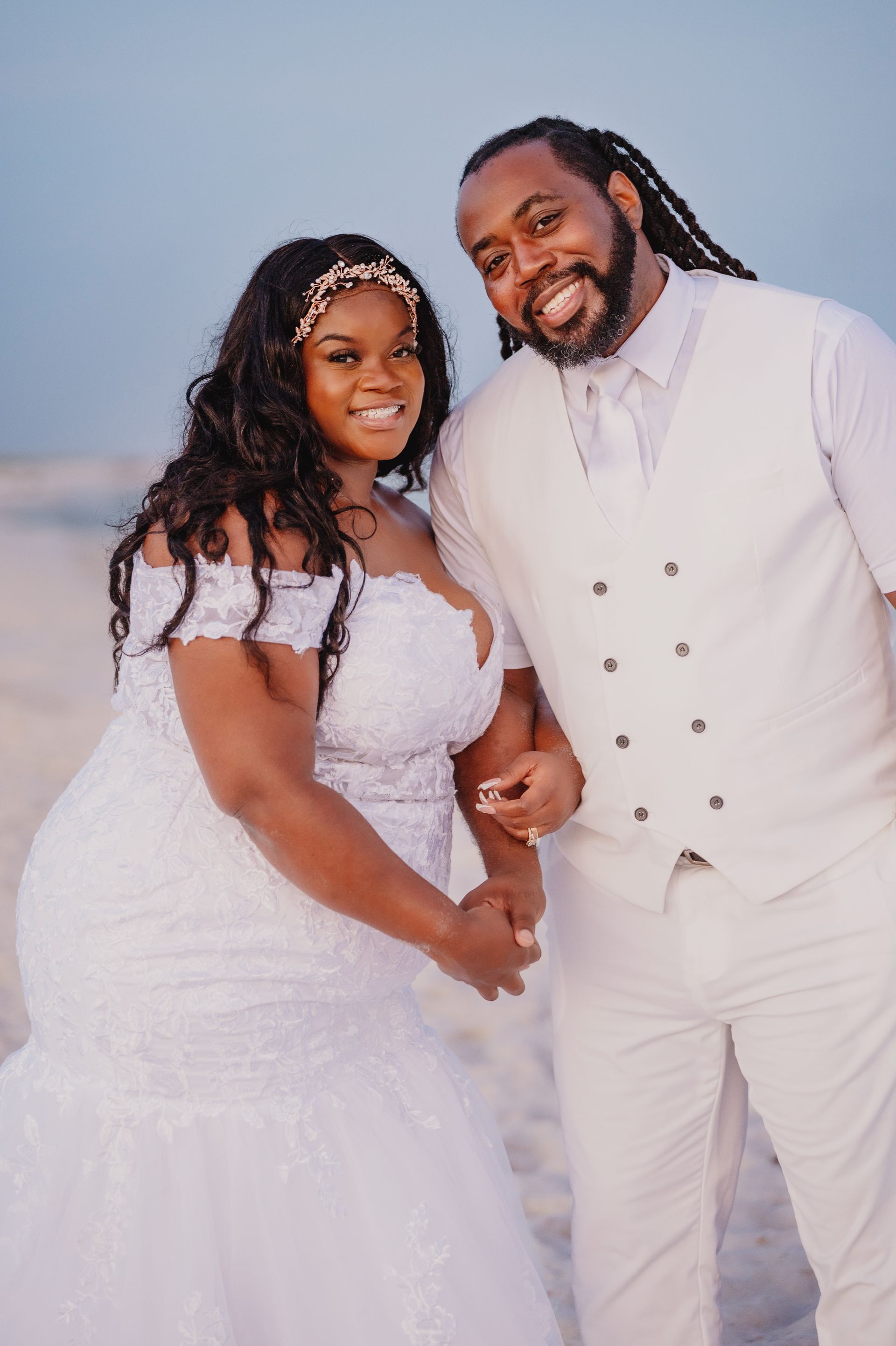 Couple in white wedding attire smiling outdoors against a soft blue sky