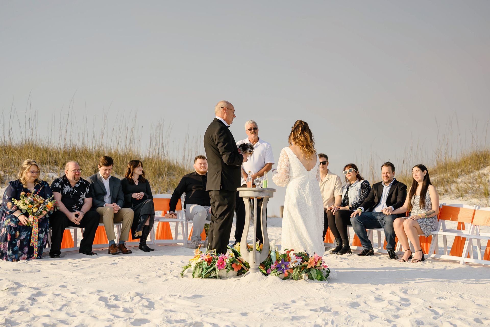 Beach wedding ceremony with bride and groom facing an officiant, guests seated nearby.