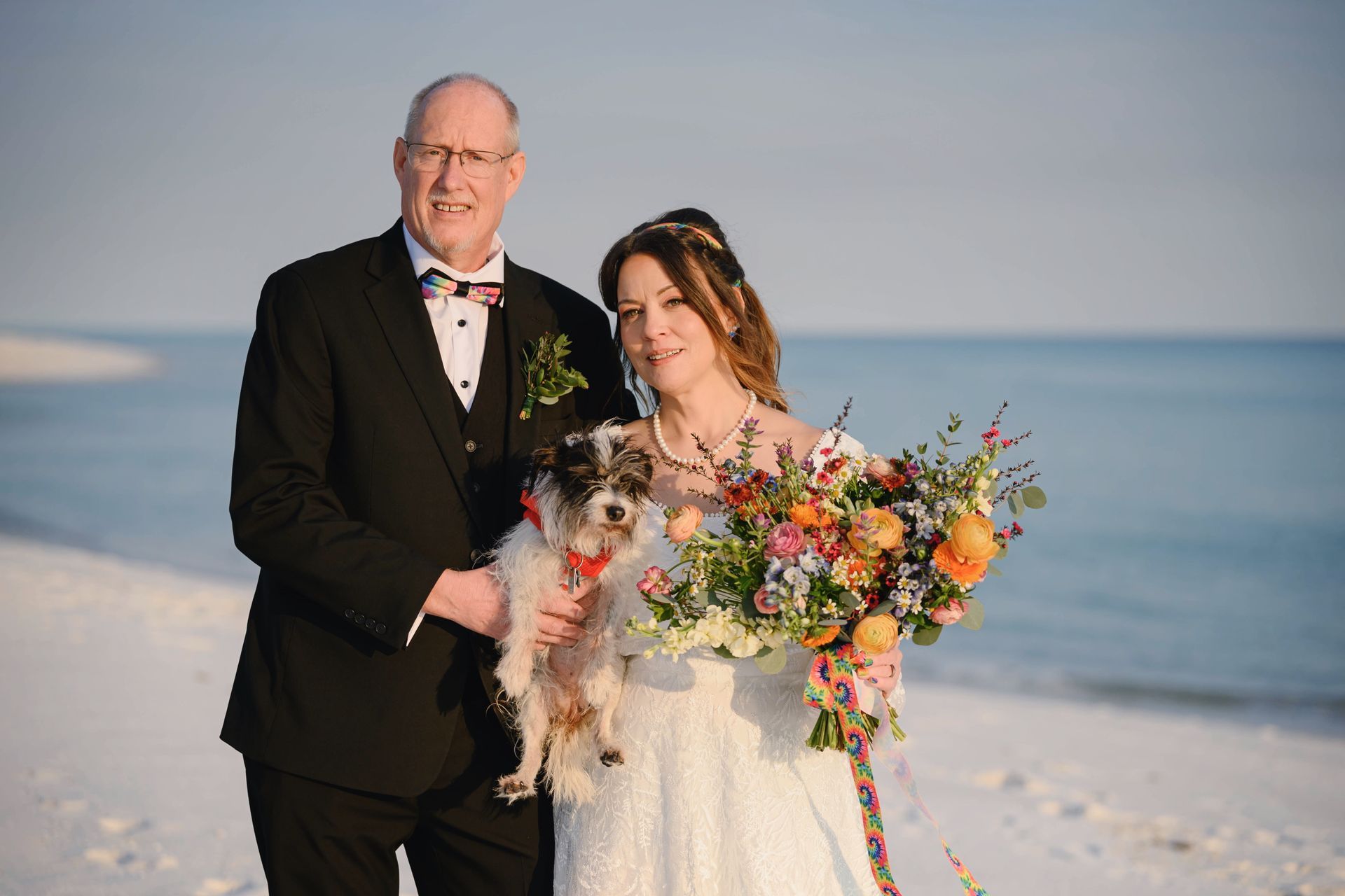 Couple in formal wear on a snowy beach, holding a small dog and colorful bouquet by the sea
