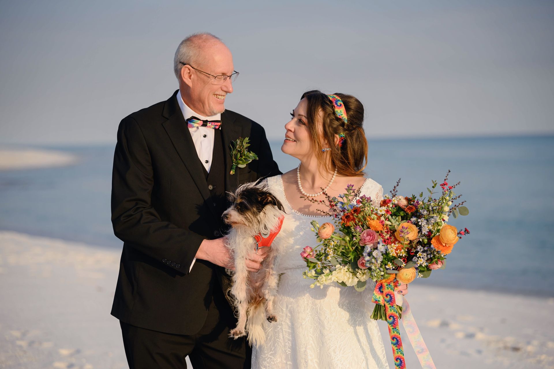 Bride and older man in formalwear holding a small dog on a beach, bride with colorful bouquet