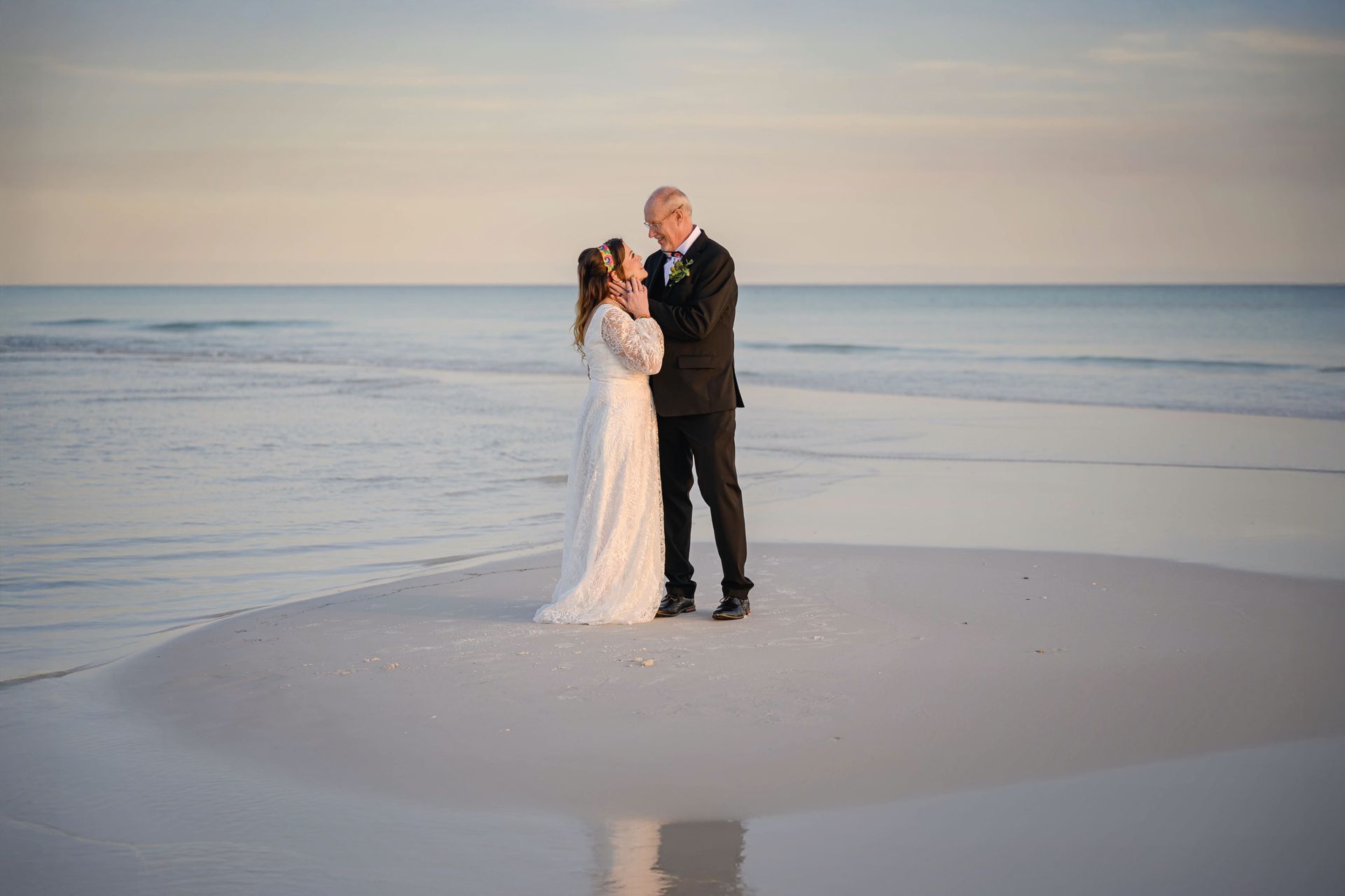 Couple in formal attire standing on a beach at sunset, reflected in shallow water.