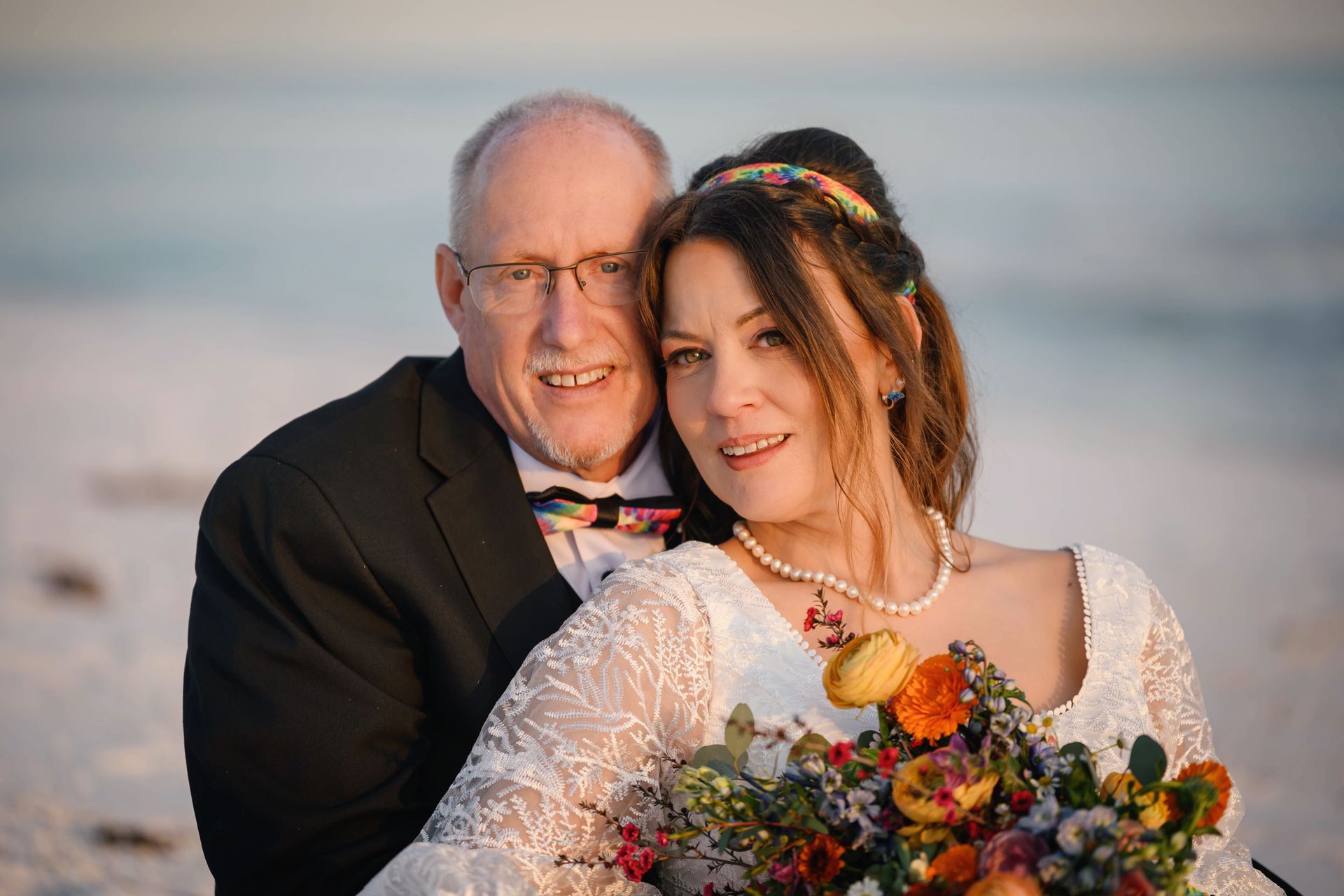 A couple in formal attire pose on a beach at sunset, smiling and holding a colorful bouquet.