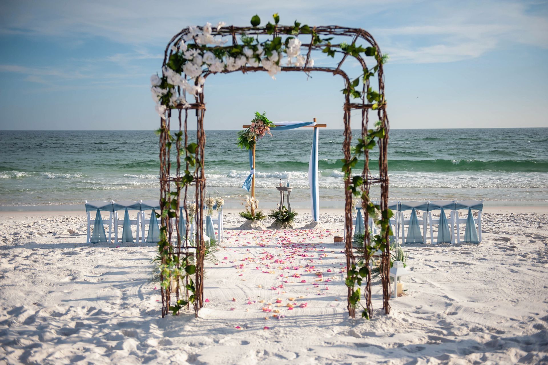 Flower-decorated wedding arch on a beach with white chairs and the ocean in the background