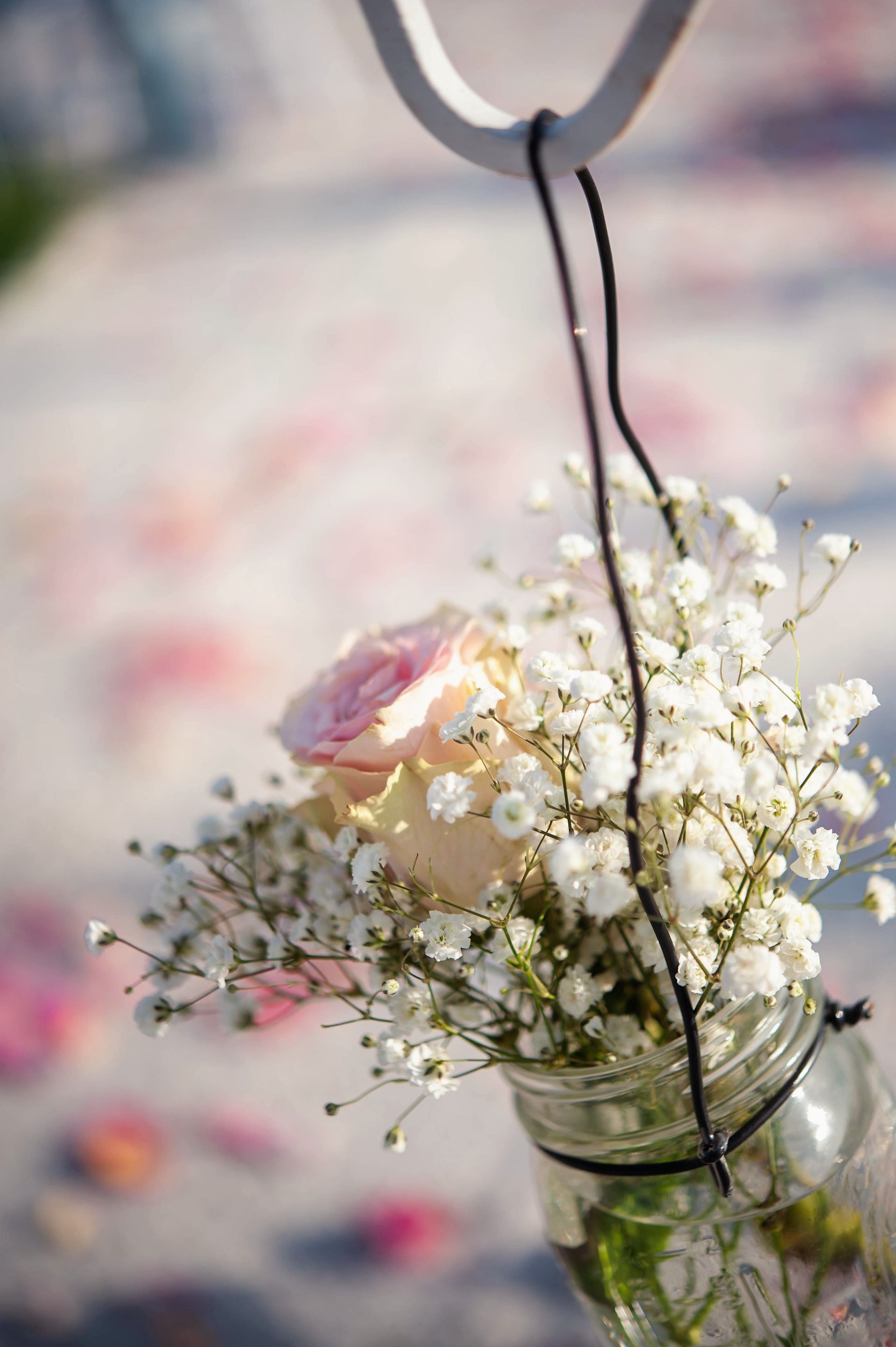 A mason jar bouquet of white baby's breath and a pink rose hanging outdoors on a blurred background