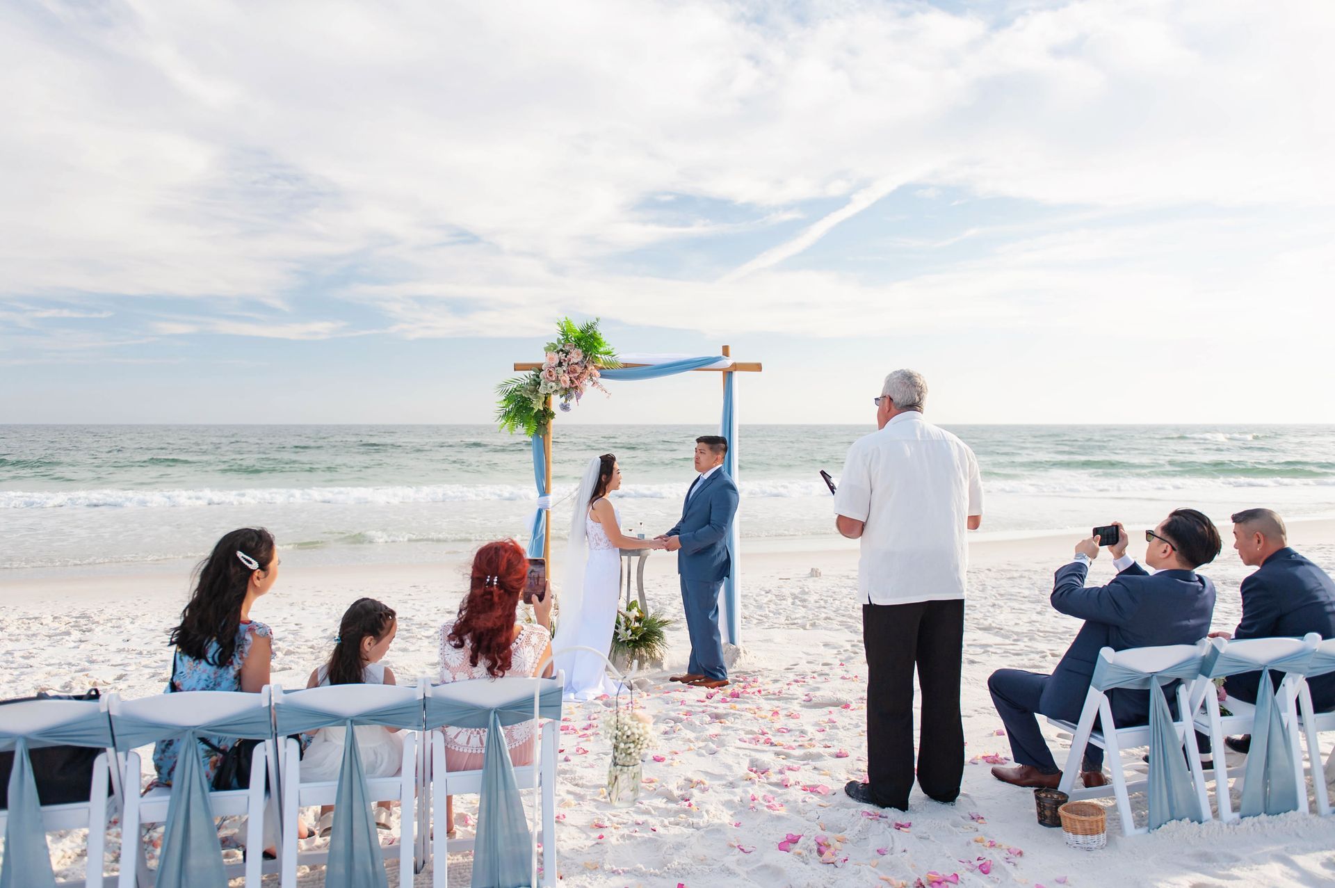 Beach wedding ceremony under a floral arch, with guests seated on white chairs by the ocean.