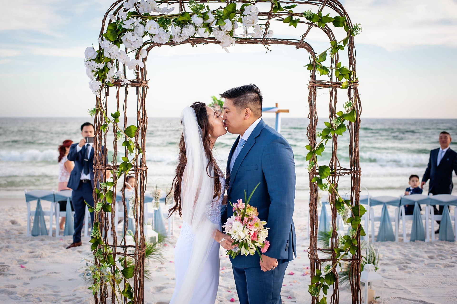 Bride and groom kiss beneath a flower-covered arch on a beach during their wedding ceremony.
