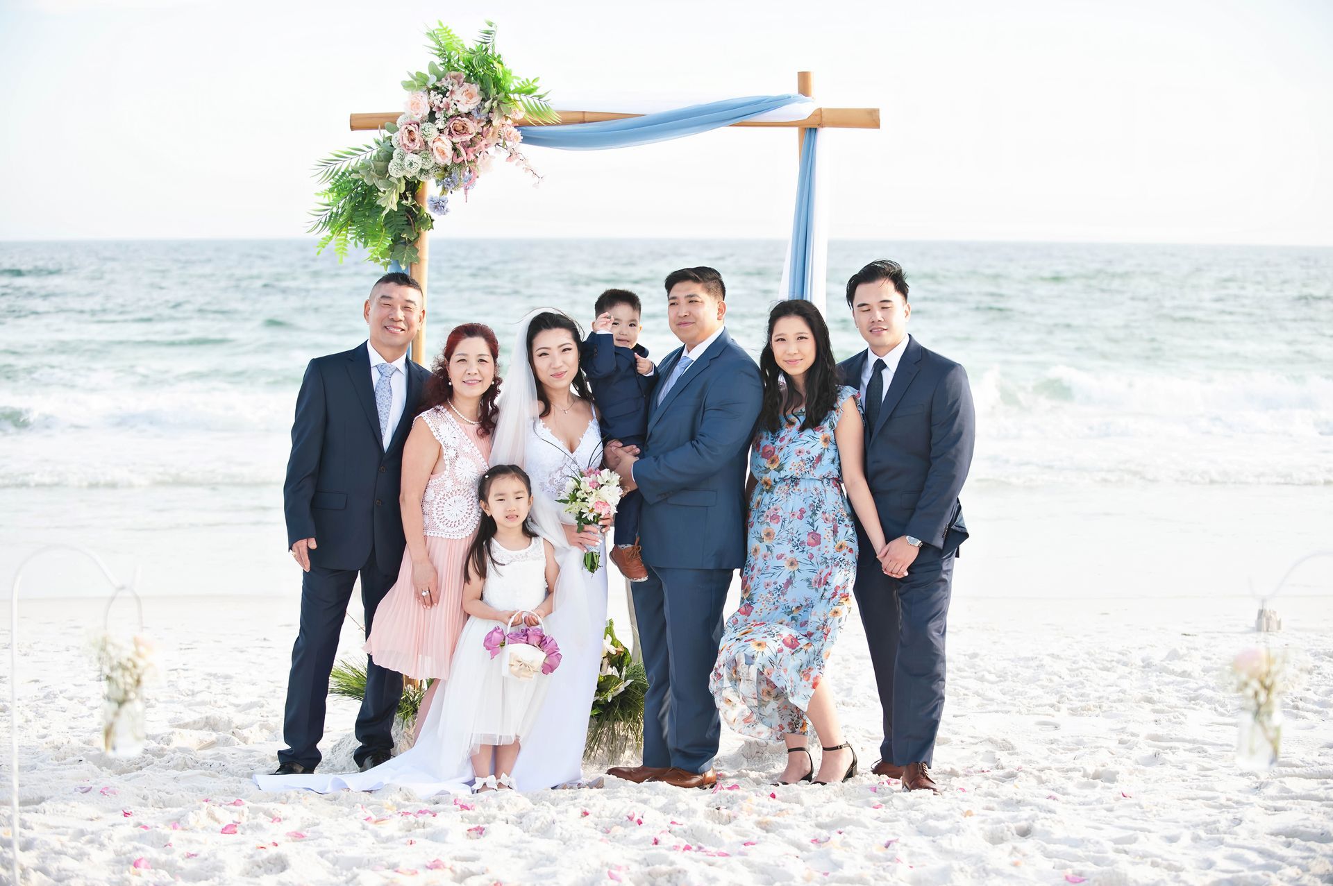 Group in formal attire posing under a floral arch on a bright beach with the ocean behind them