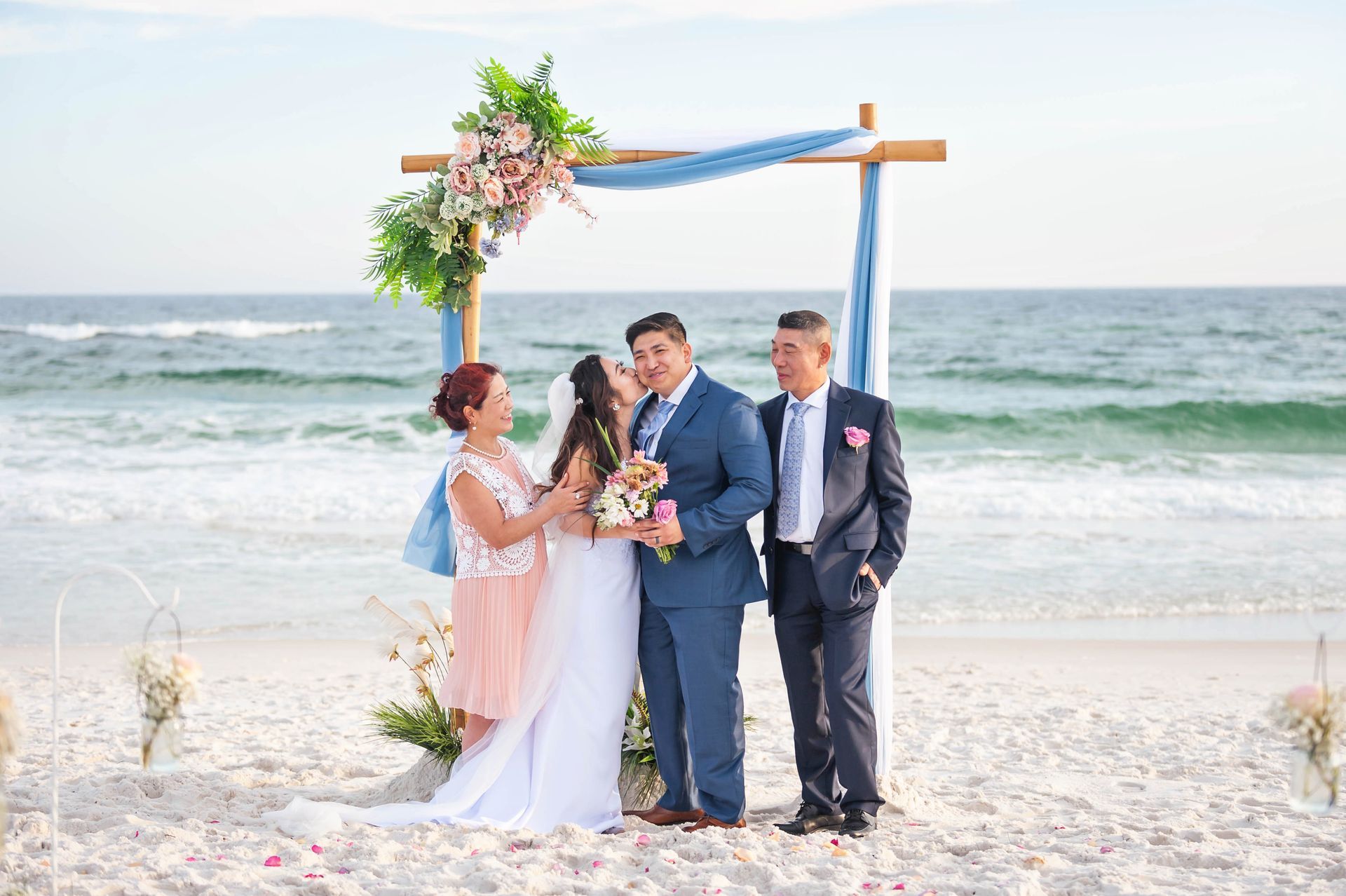 Wedding couple and guest standing under a floral arch on a sunny beach by the ocean