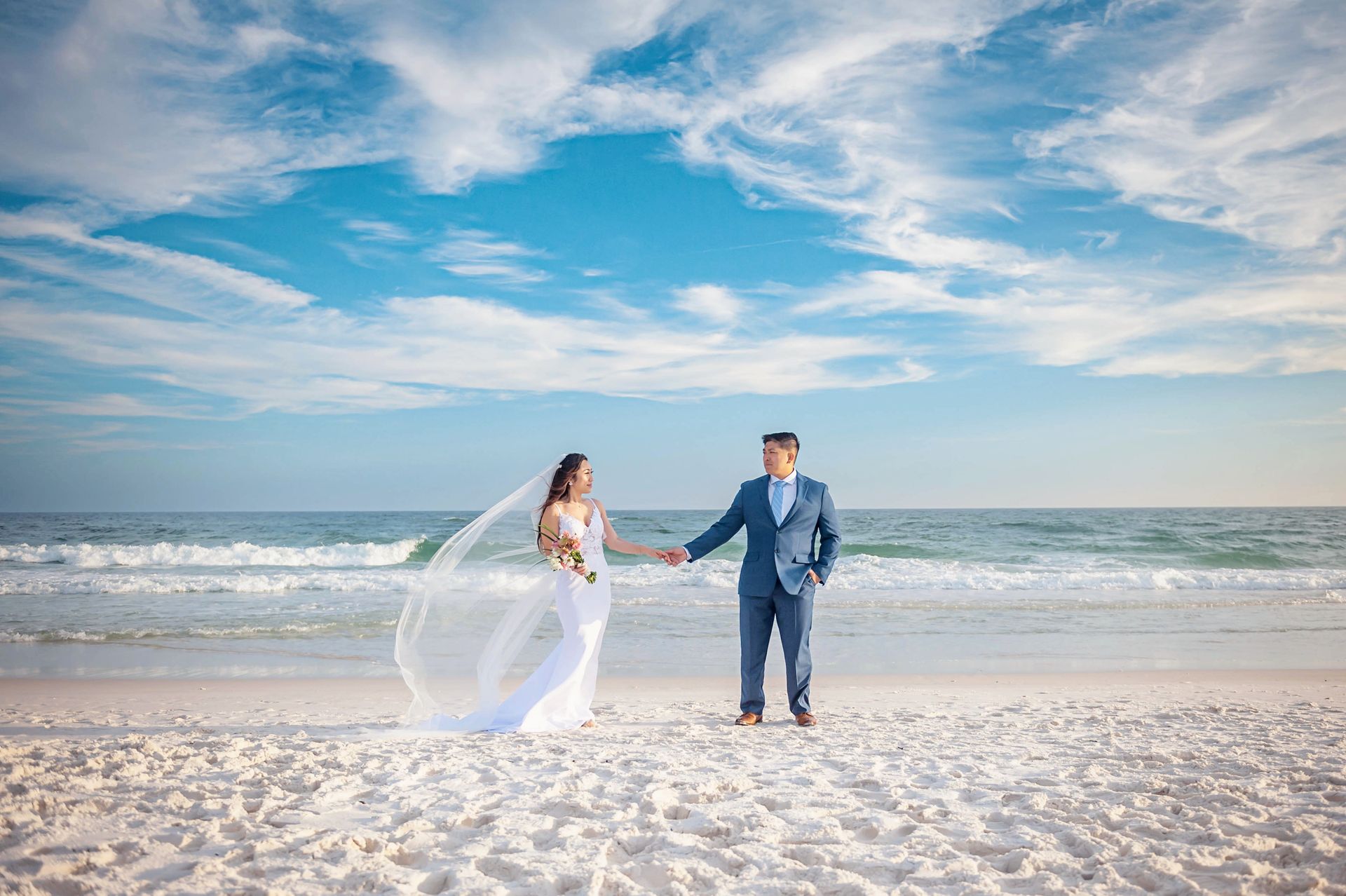Couple in wedding attire holding hands on a bright beach under a blue sky