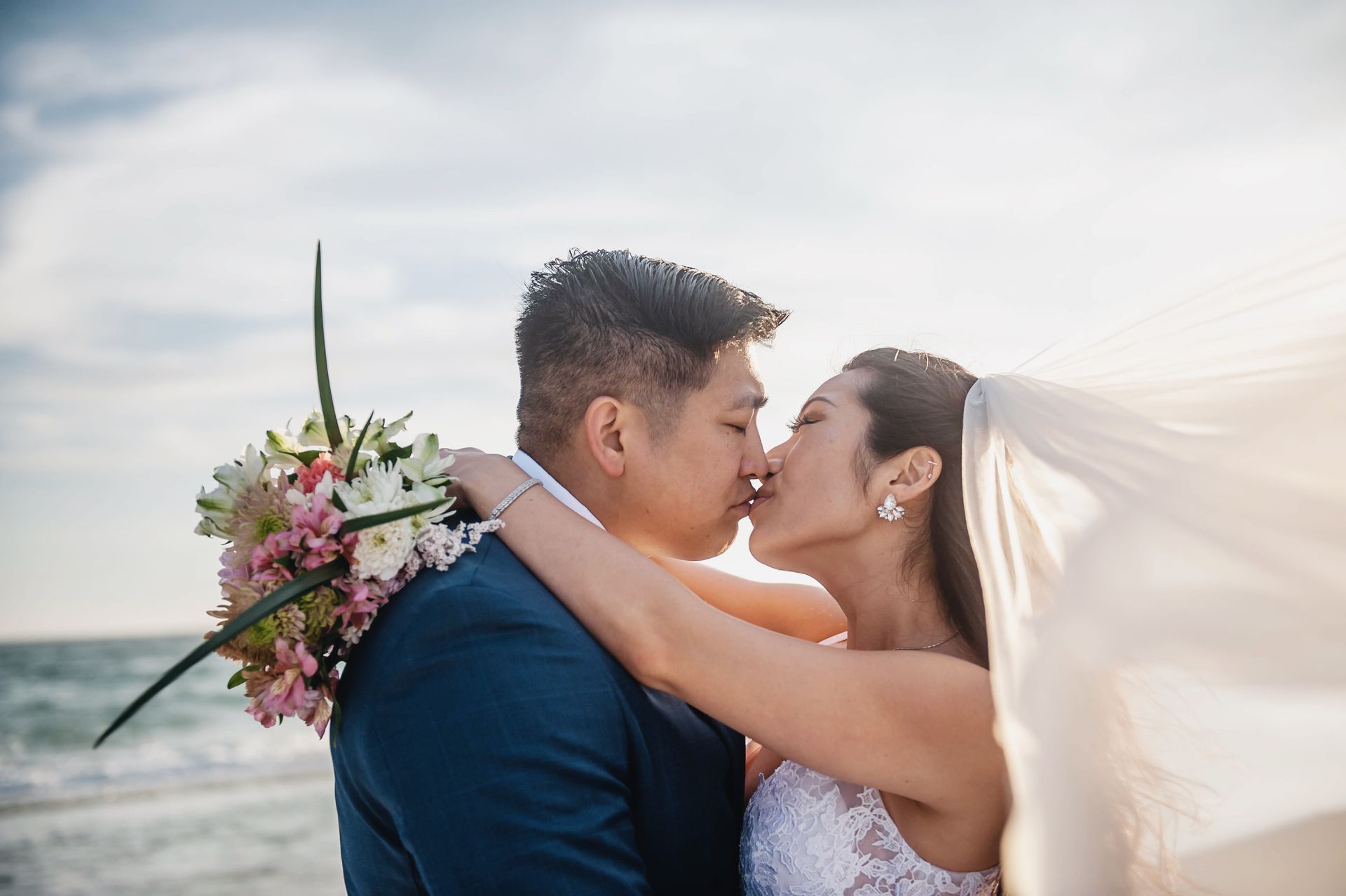 Bride and groom embracing and kissing on the beach, with the veil blowing in the wind