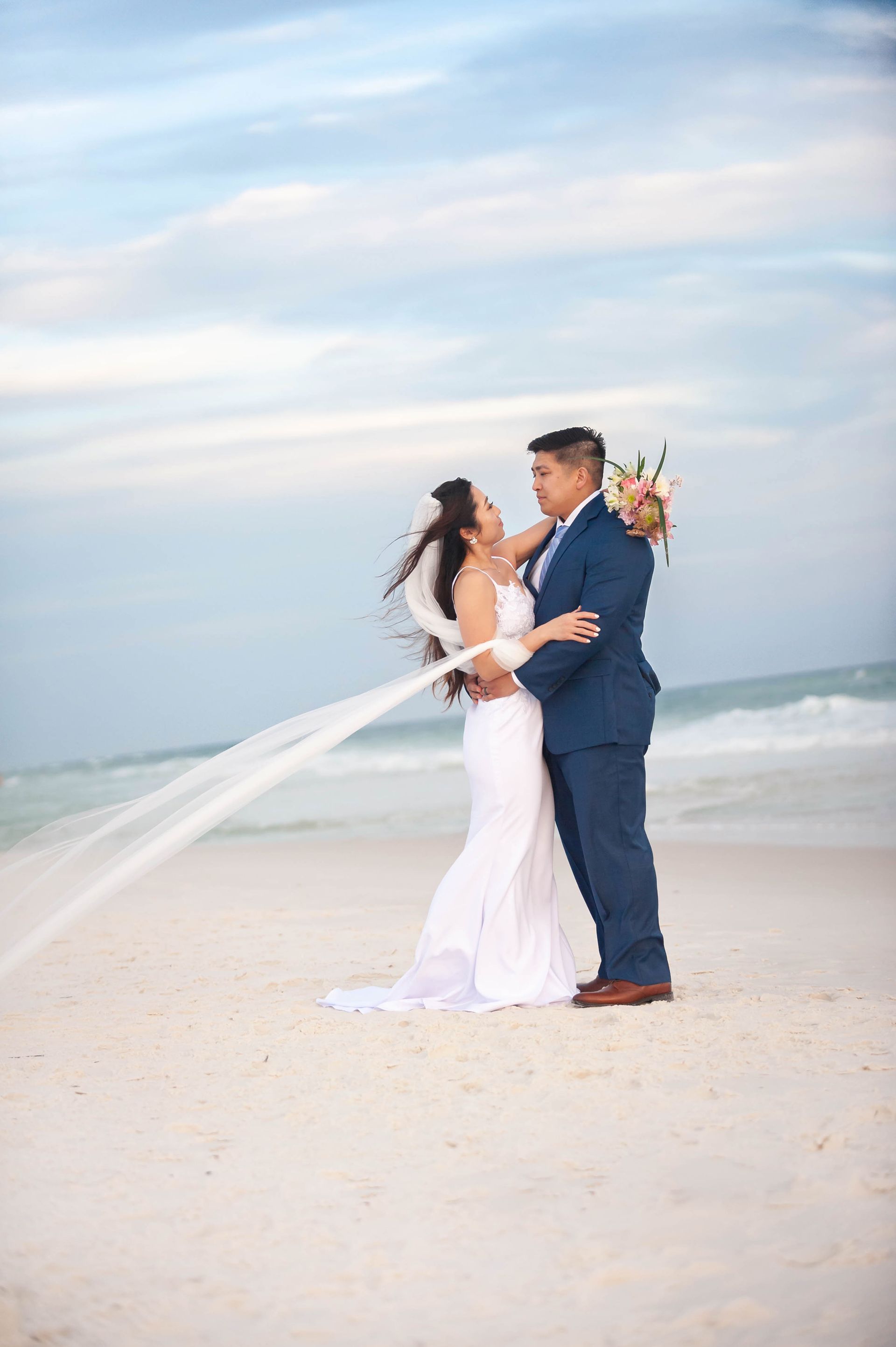 Bride and groom embracing on a beach with ocean waves and a flowing veil