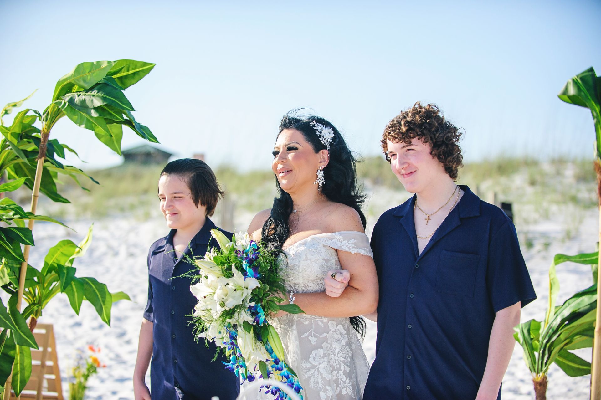 Three people walking outdoors in bright sunlight, with the center person holding a bouquet.