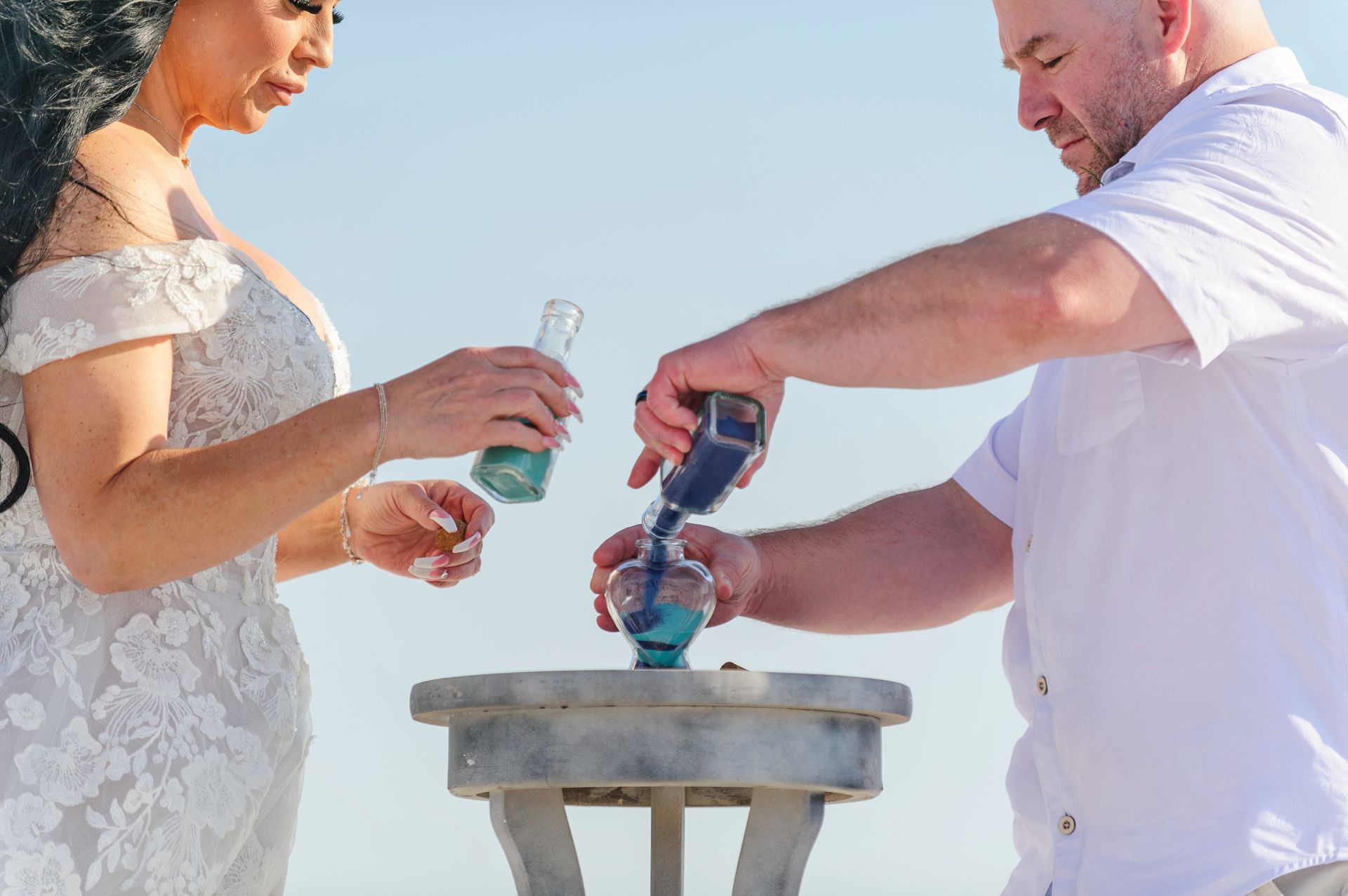Bride and groom pour drinks together at a small outdoor table in bright daylight