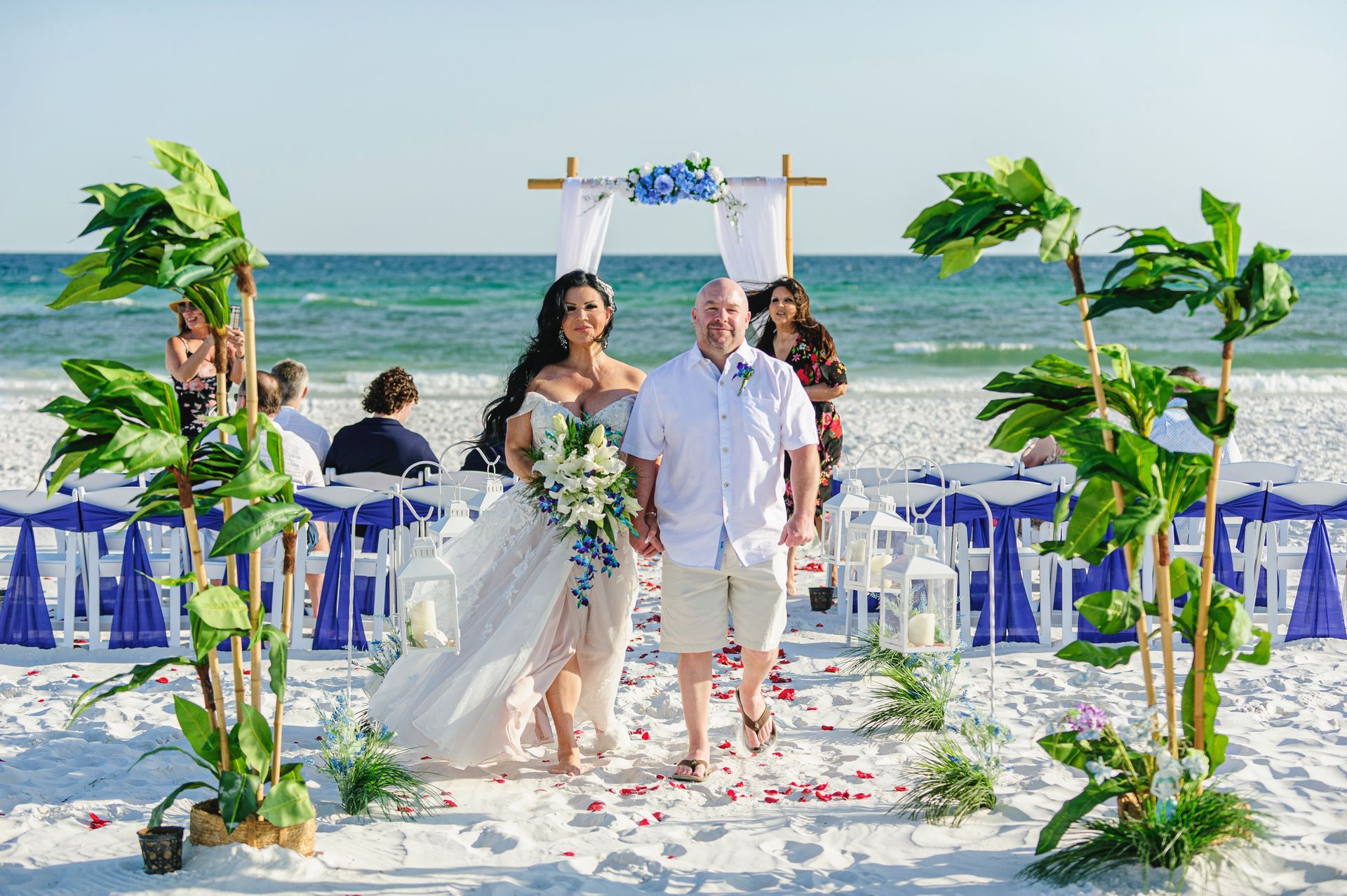 Beach wedding couple walking down an aisle under a white arch with blue chairs and palm decorations