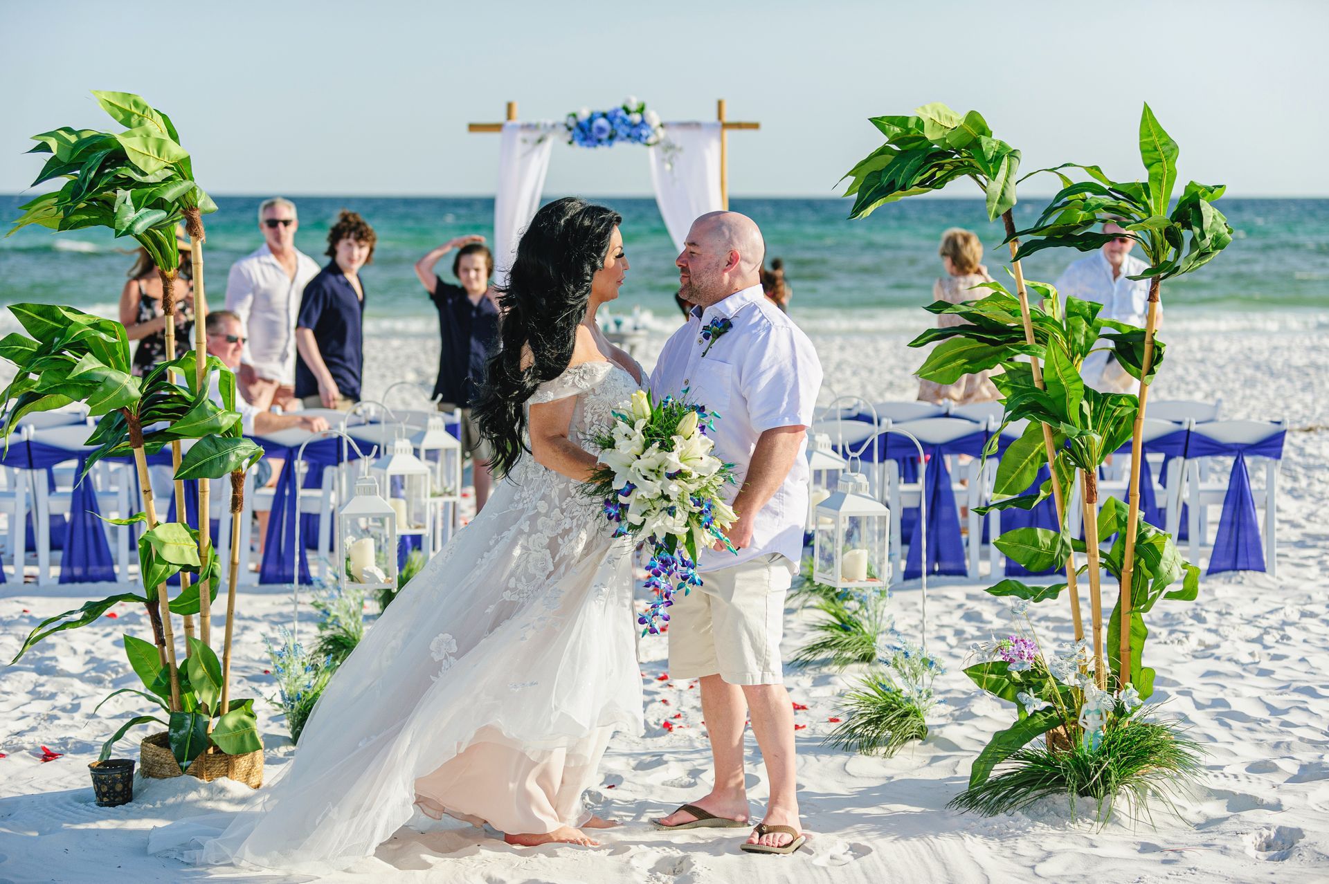 Beach wedding ceremony with bride and groom under floral arch, blue chairs on the sand, ocean behind them