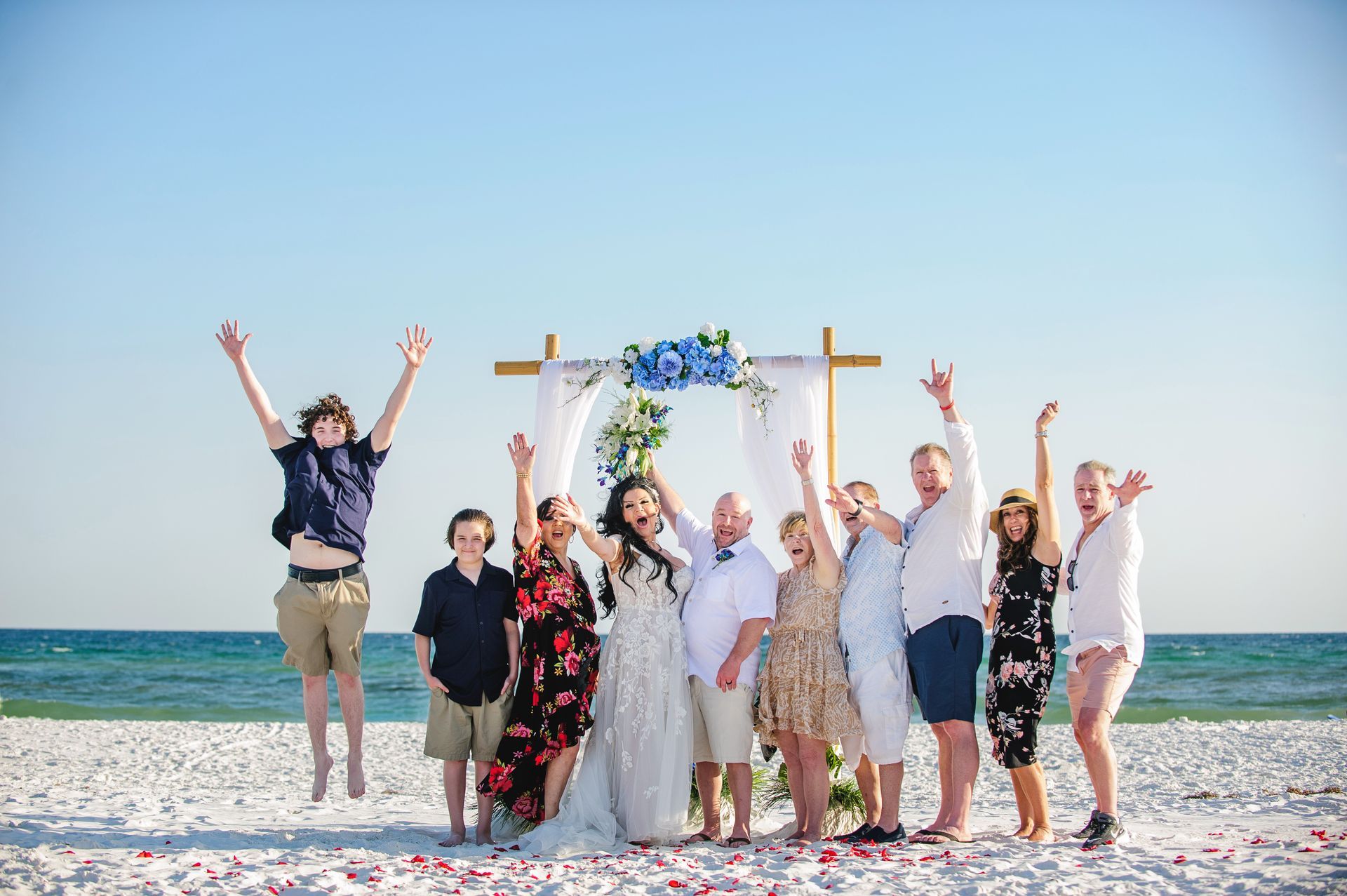 Group of people celebrating on a beach under a floral arch, with some jumping and raising their arms.